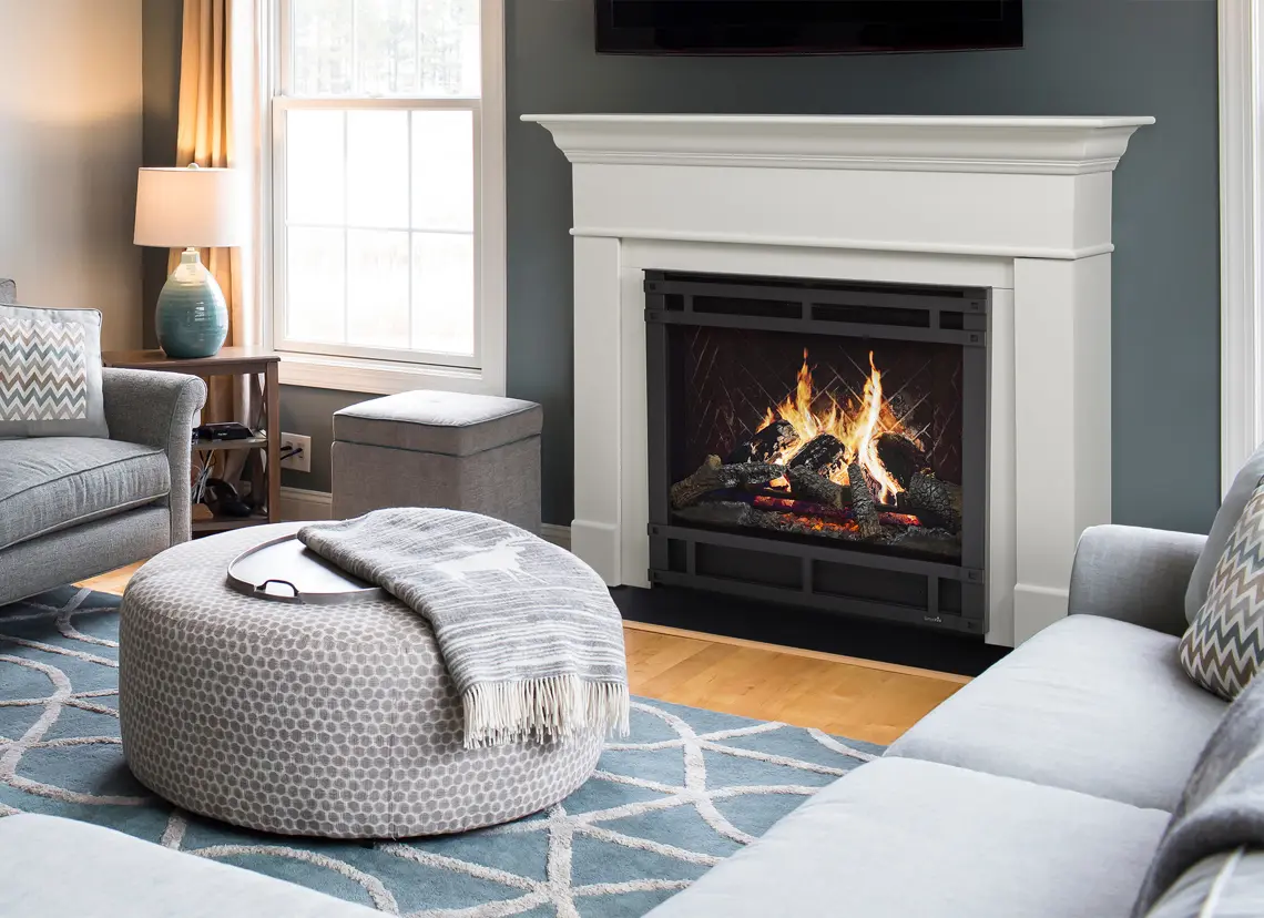 A cozy living room with a lit fireplace, a gray couch, a patterned ottoman with a tray and blanket, a side table with a lamp, and a large window with curtains.