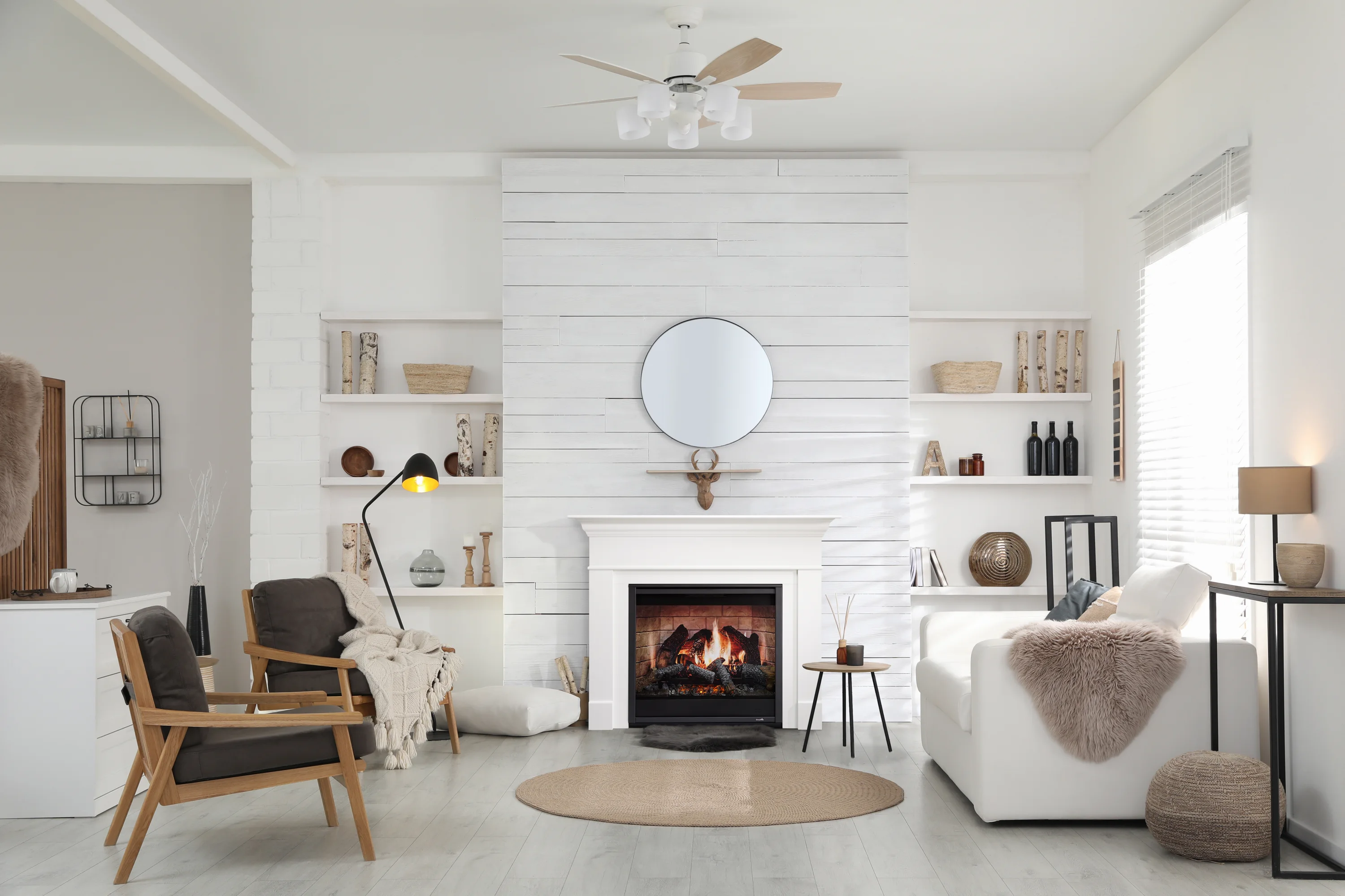 Modern living room with a fireplace, two armchairs, and a white sofa. The room features white walls and shelves with decorative items, a round mirror above the fireplace, and a ceiling fan.