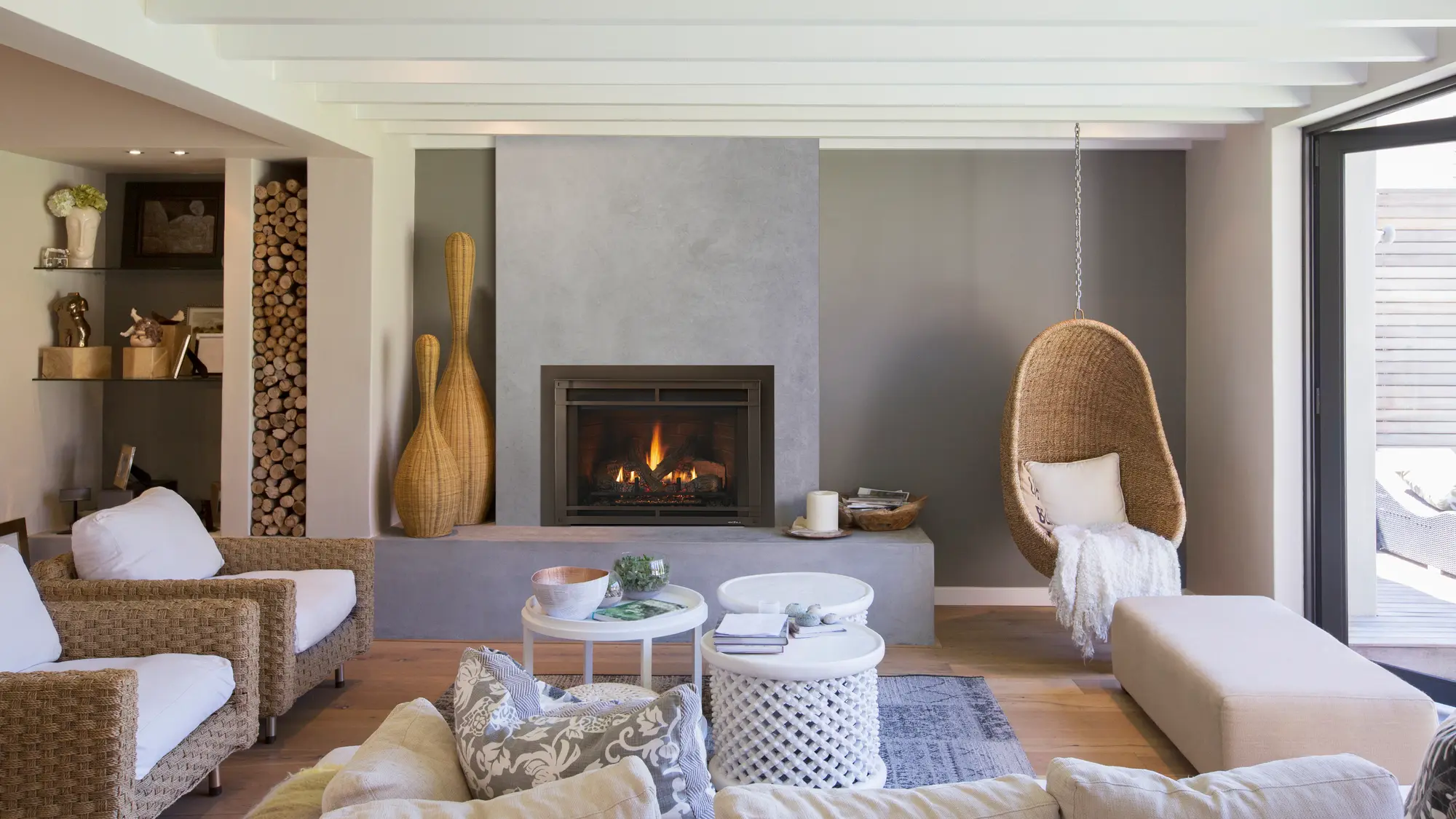 Modern living room with a lit fireplace, wicker chairs, a hanging wicker swing chair with a pillow and blanket, and two white tables. Beige walls, wooden floor, and a wood storage niche.
