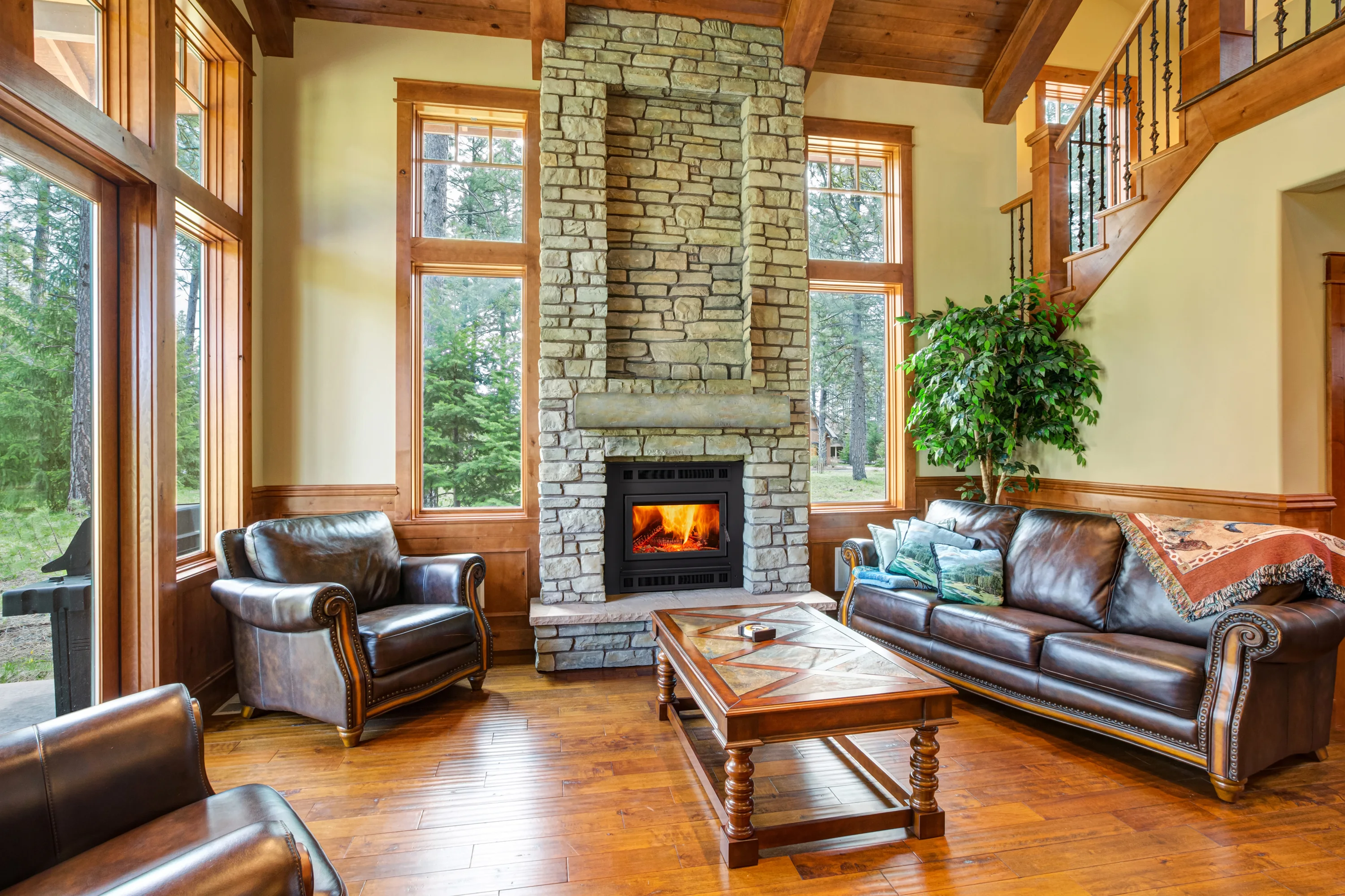 A cozy living room with high ceilings, large windows, a stone fireplace with a lit fire, brown leather sofas, an armchair, a wooden coffee table, and a staircase in the background.