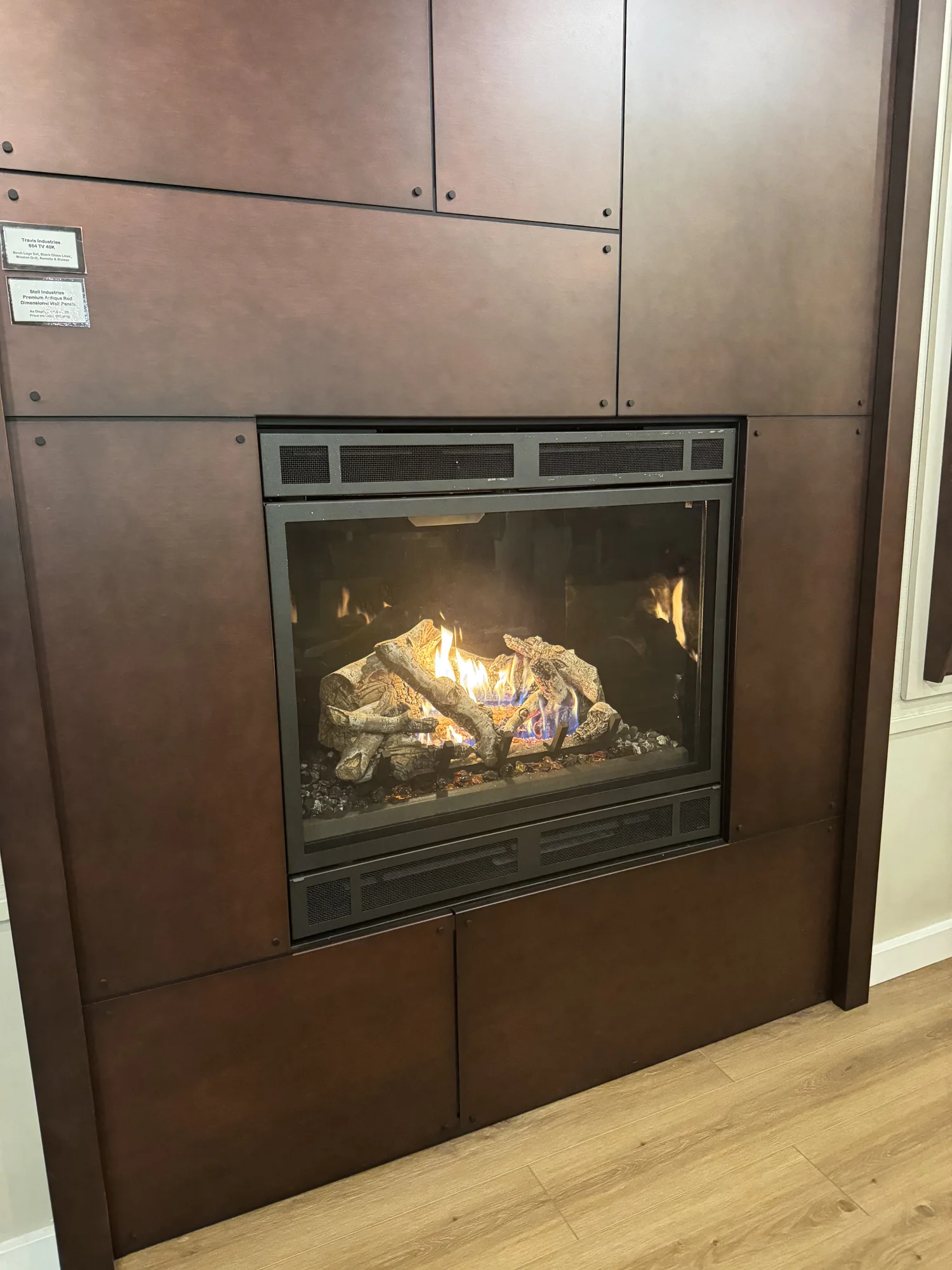 Gas fireplace with glowing ceramic logs and blue and yellow flames in a black metal-framed glass insert set into a dark brown paneled wall above light wood floor