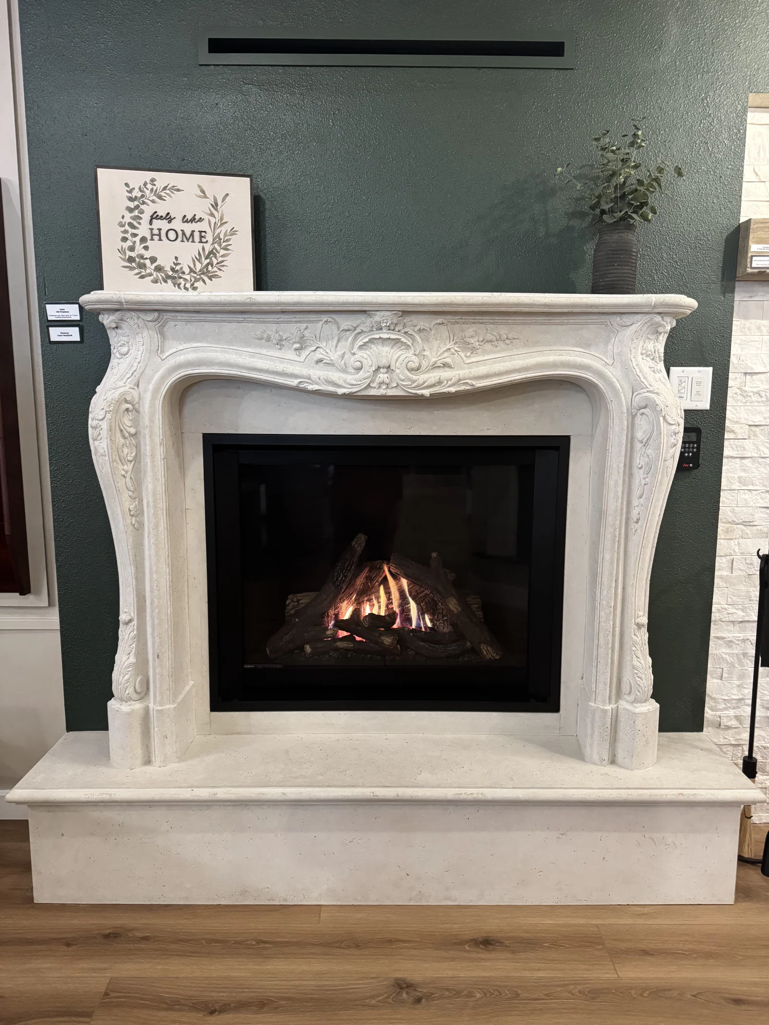 Ornate white stone fireplace mantel with a burning gas fire, dark green accent wall, framed feels like HOME sign on left and a tall vase of greenery on right