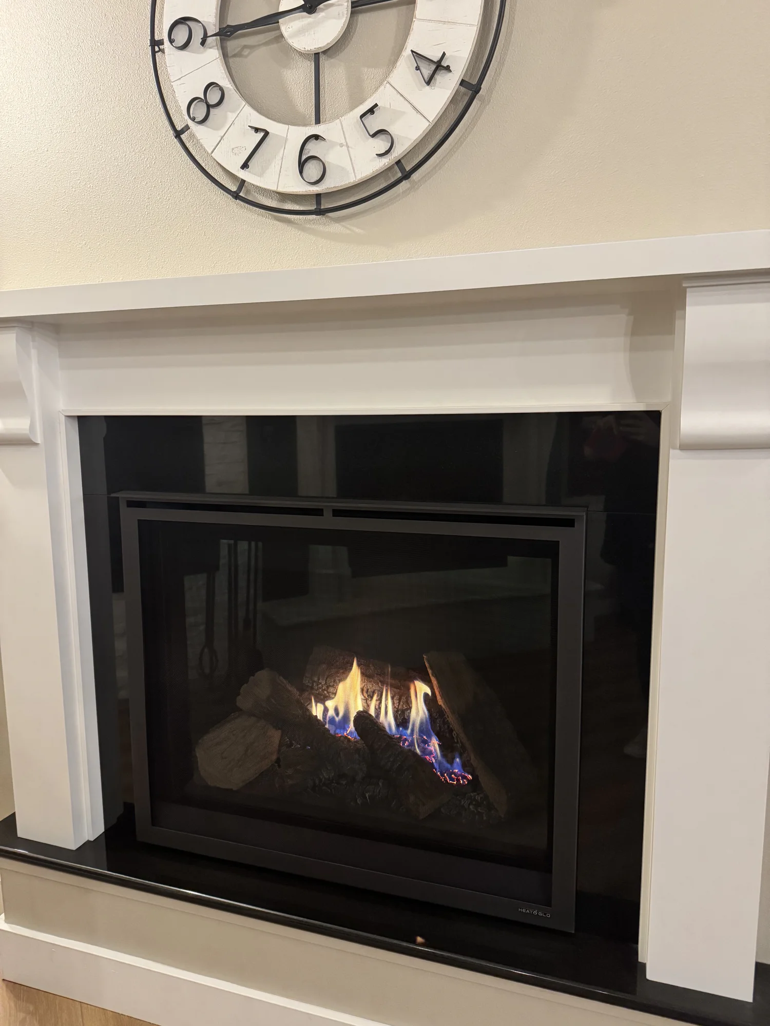 Lit gas fireplace with black glass door and logs under a white mantel, partial round wall clock above
