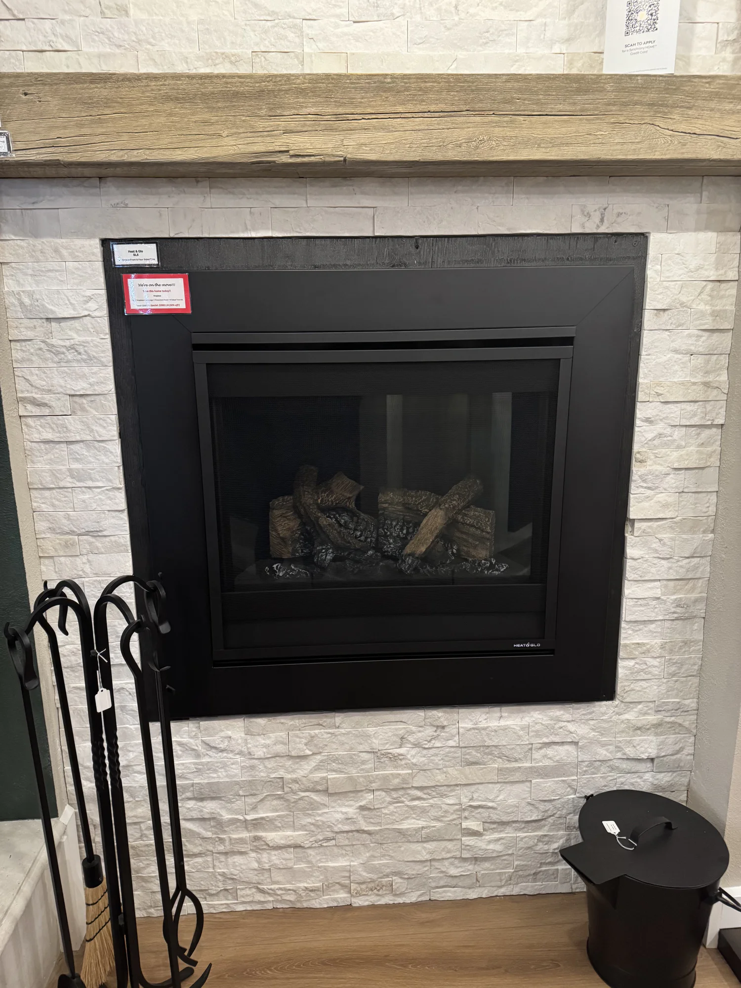 Black-framed gas fireplace with ceramic logs, white stacked stone surround and reclaimed wood mantel, iron fireplace tools at left and black ash bucket at right