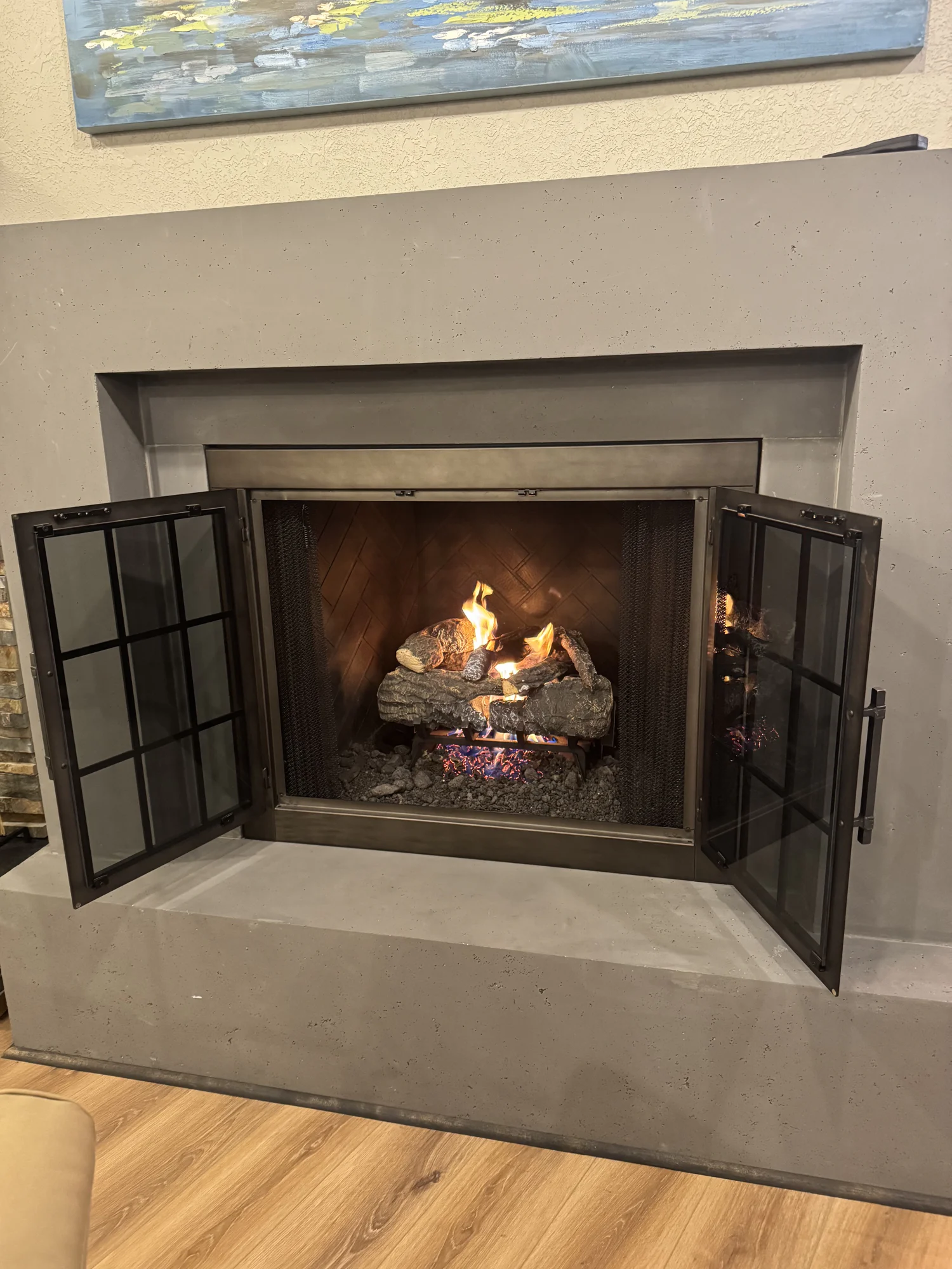 Modern built-in gas fireplace with metal-framed glass doors open, burning ceramic logs and glowing embers, concrete surround, wood floor and painting visible above.