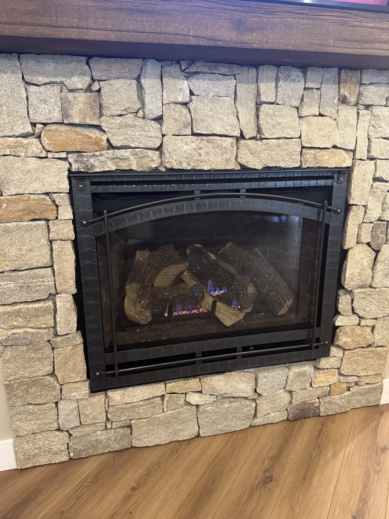 Stone-faced gas fireplace with black metal frame, visible ceramic logs and small blue flames, wooden mantel above and hardwood floor below.