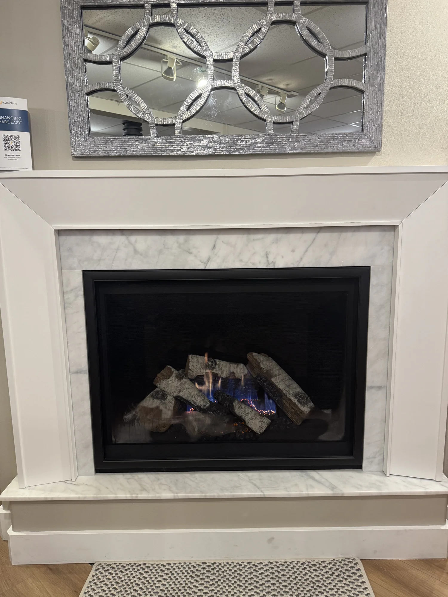 White fireplace with marble surround and black firebox showing gas logs with blue flames, and a silver decorative mirror mounted above