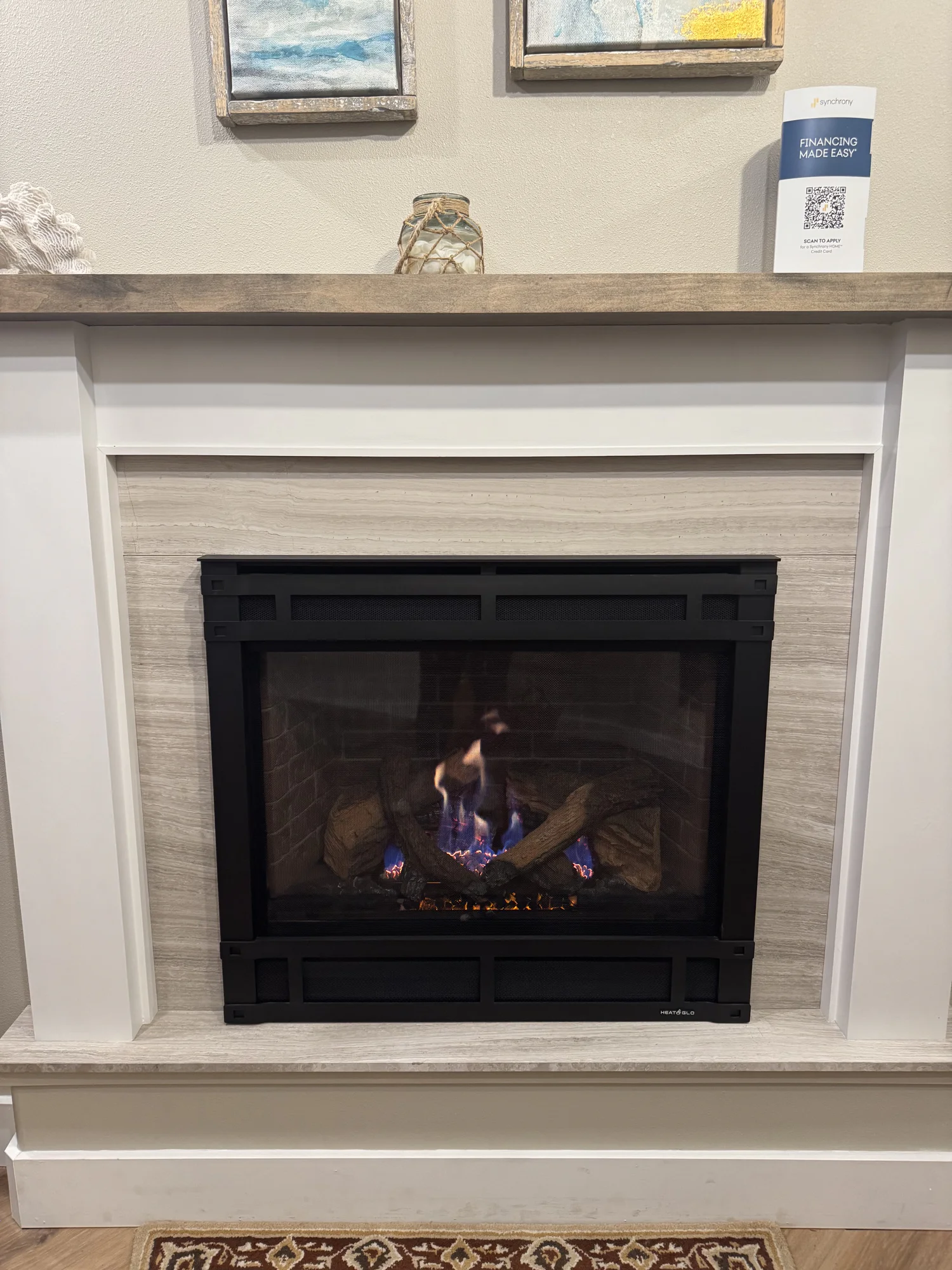 Lit gas fireplace with black metal frame set in white mantel and light wood-look surround; mantle holds a glass jar wrapped in rope and a small financing brochure.
