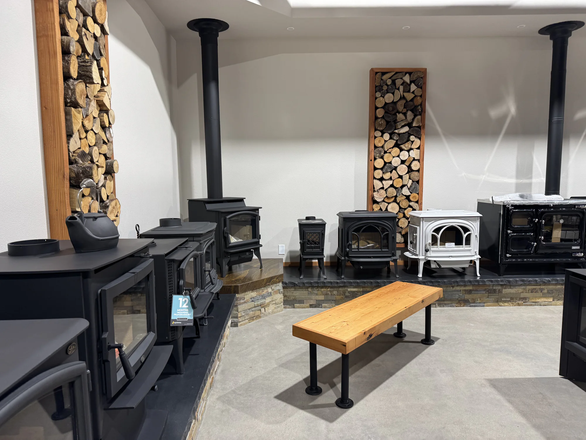Showroom display of various wood-burning stoves with vertical stacked firewood and a wooden bench.