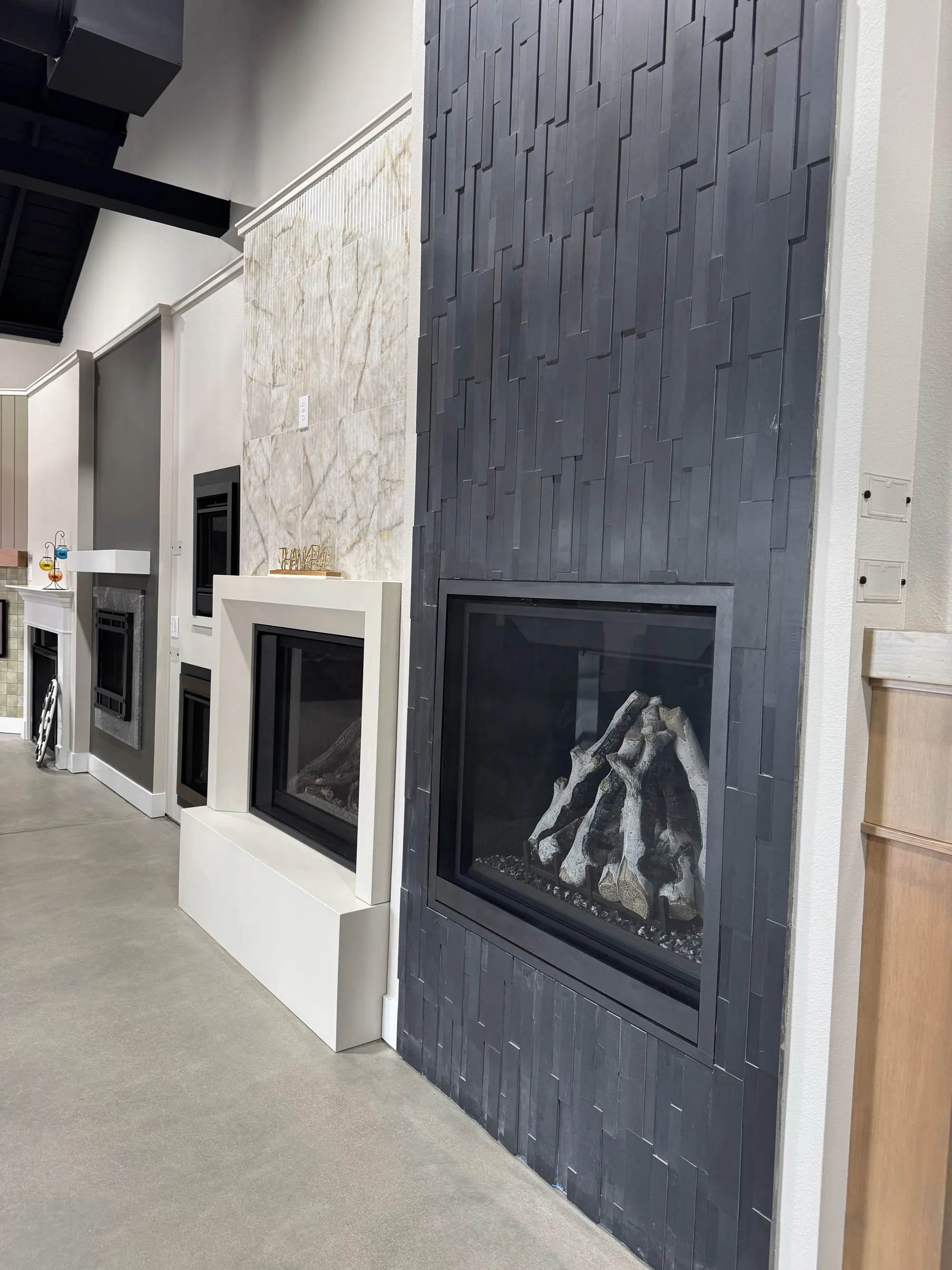 Showroom display of modern fireplaces, dark textured tile fireplace in foreground with white marble surrounds behind