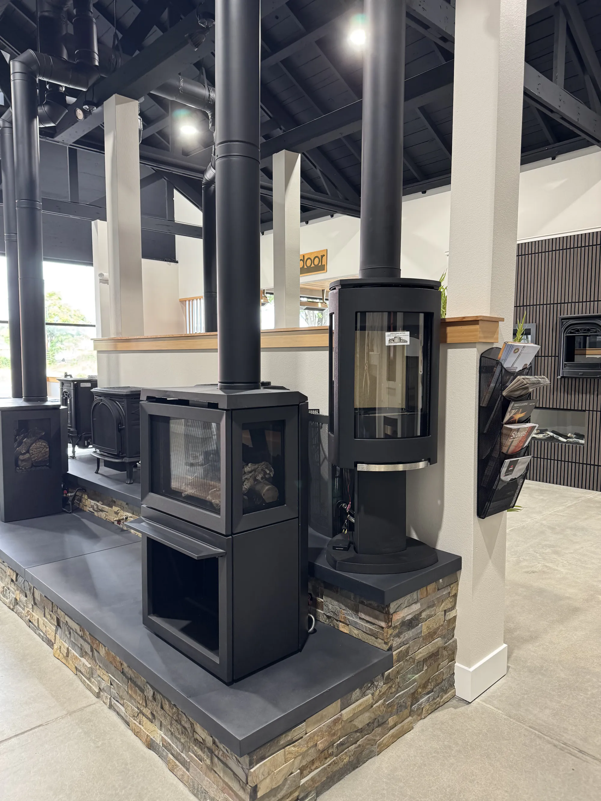 Showroom display of black freestanding wood stoves with tall chimneys on a raised stone platform