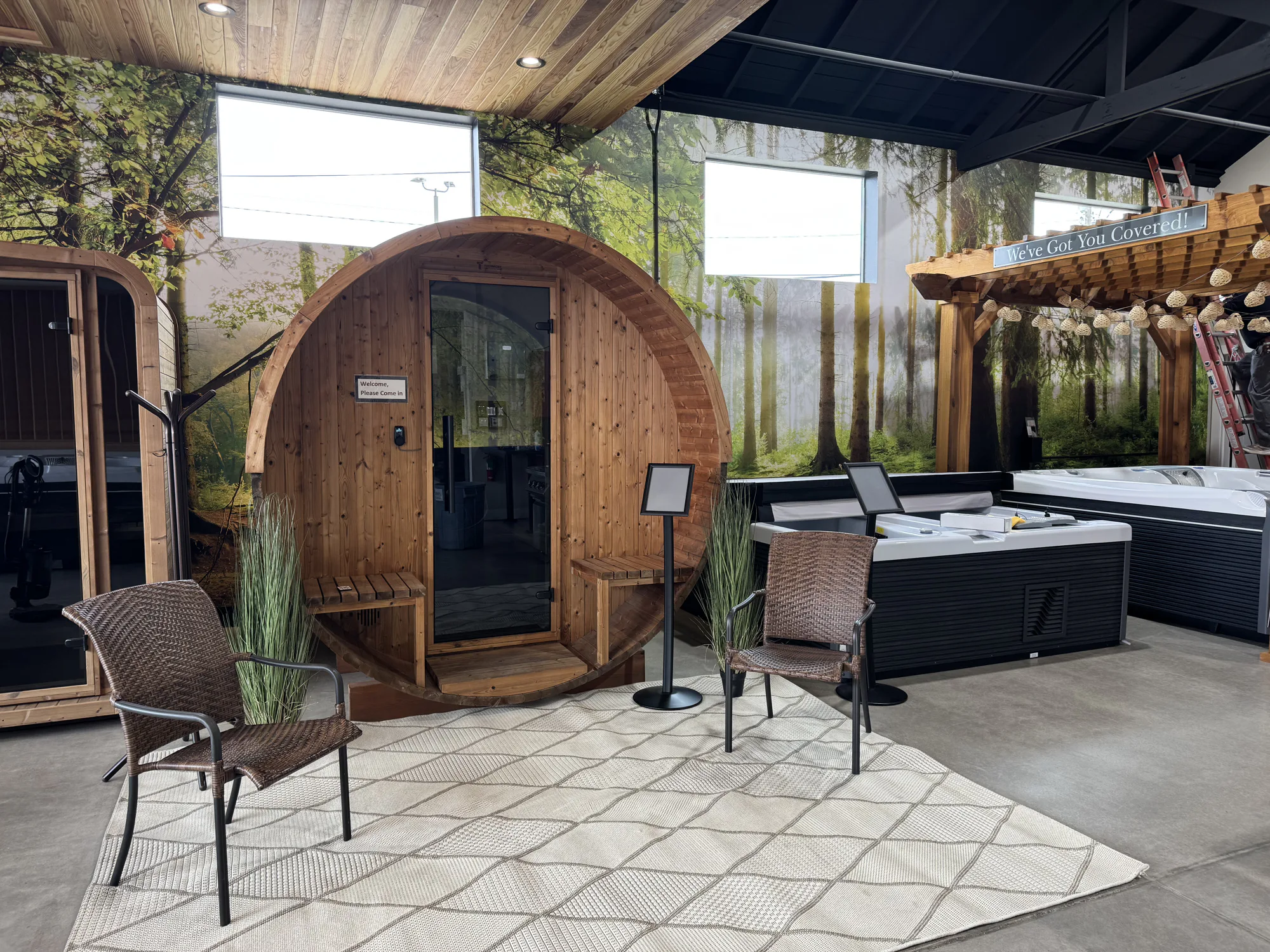 Indoor showroom with wooden barrel sauna, pergola, hot tubs and wicker chairs against a forest mural backdrop