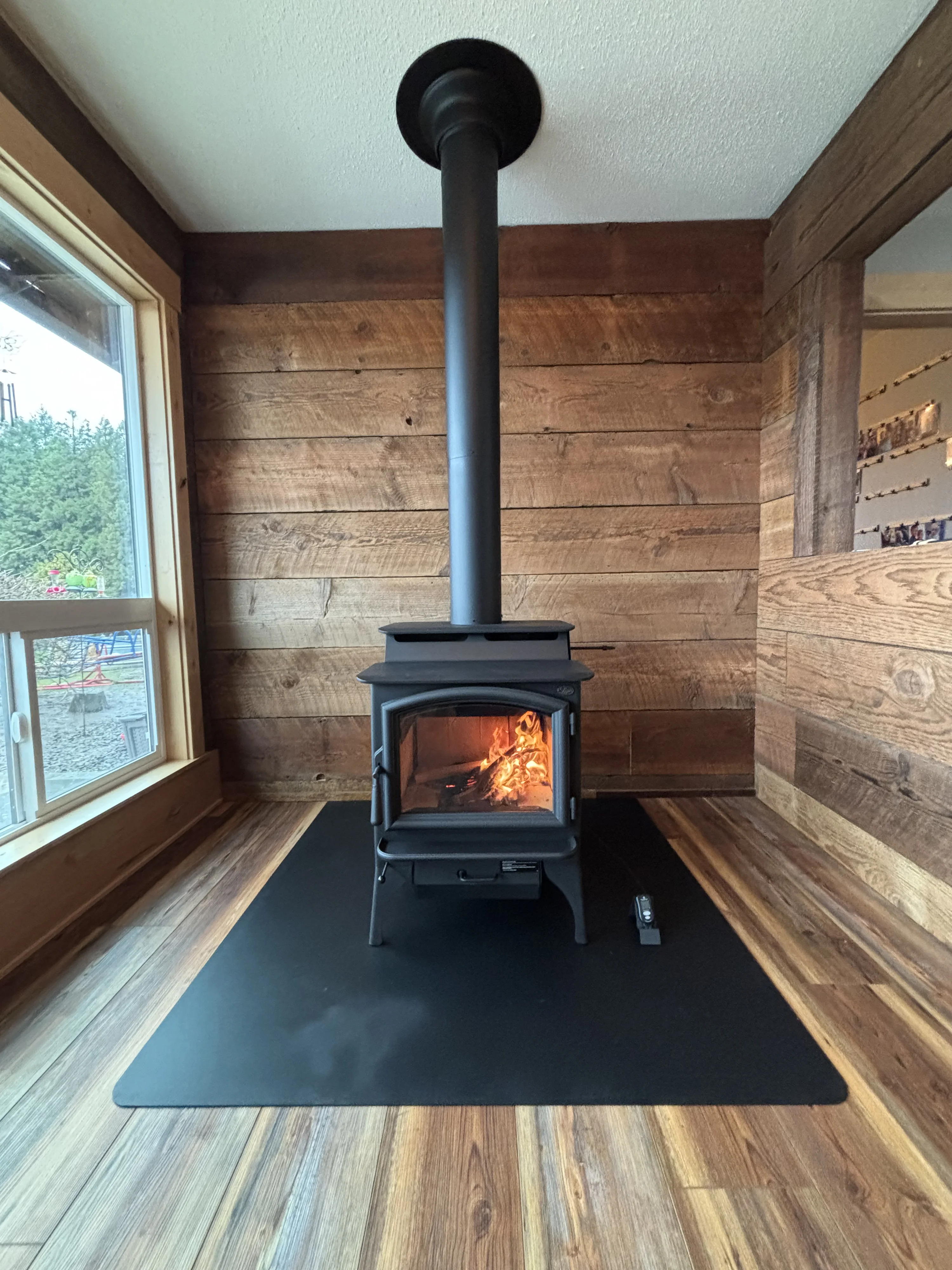 A wood stove with a burning fire is located in a corner of a room with wooden walls and a window overlooking a partly visible outdoor area, all set on a wooden floor with a black mat.