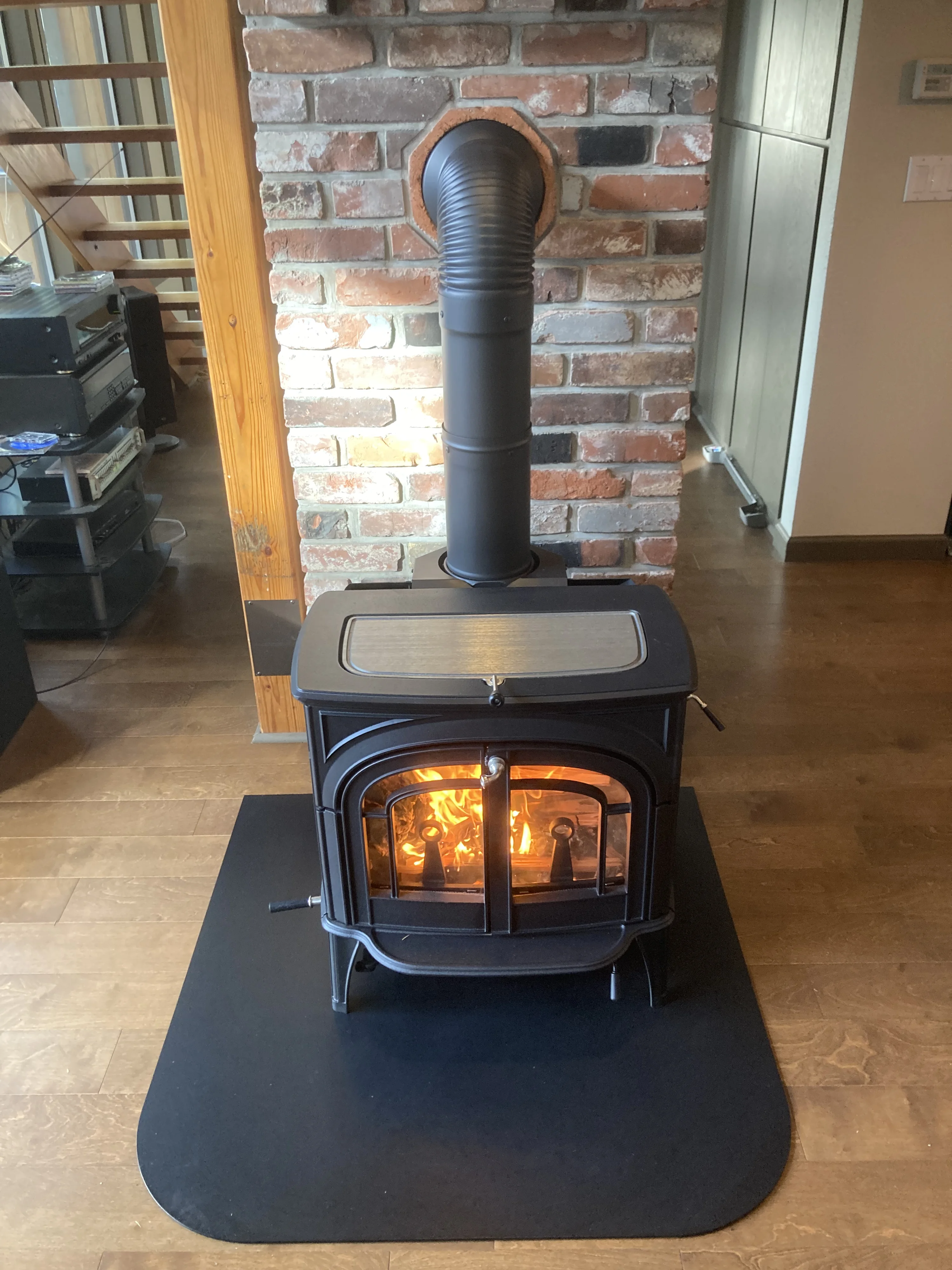 Black cast-iron wood stove with glowing fire, vertical stovepipe into a brick wall, sitting on a black hearth pad on hardwood floor