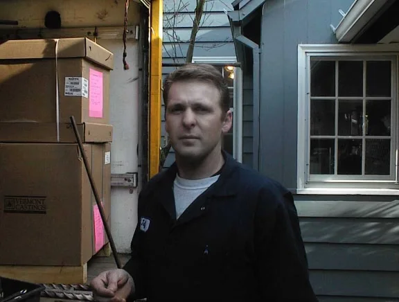 A man in a dark uniform stands outside by a house. Empty moving boxes are stacked on a dolly next to him. A window and a closed door of the house are visible in the background.