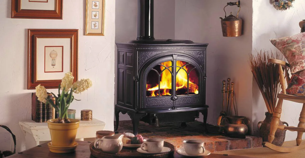 A cozy room with a black wood-burning stove surrounded by framed art on the walls, a yellow potted plant on a white table, and a tea set on a wooden table in the foreground.