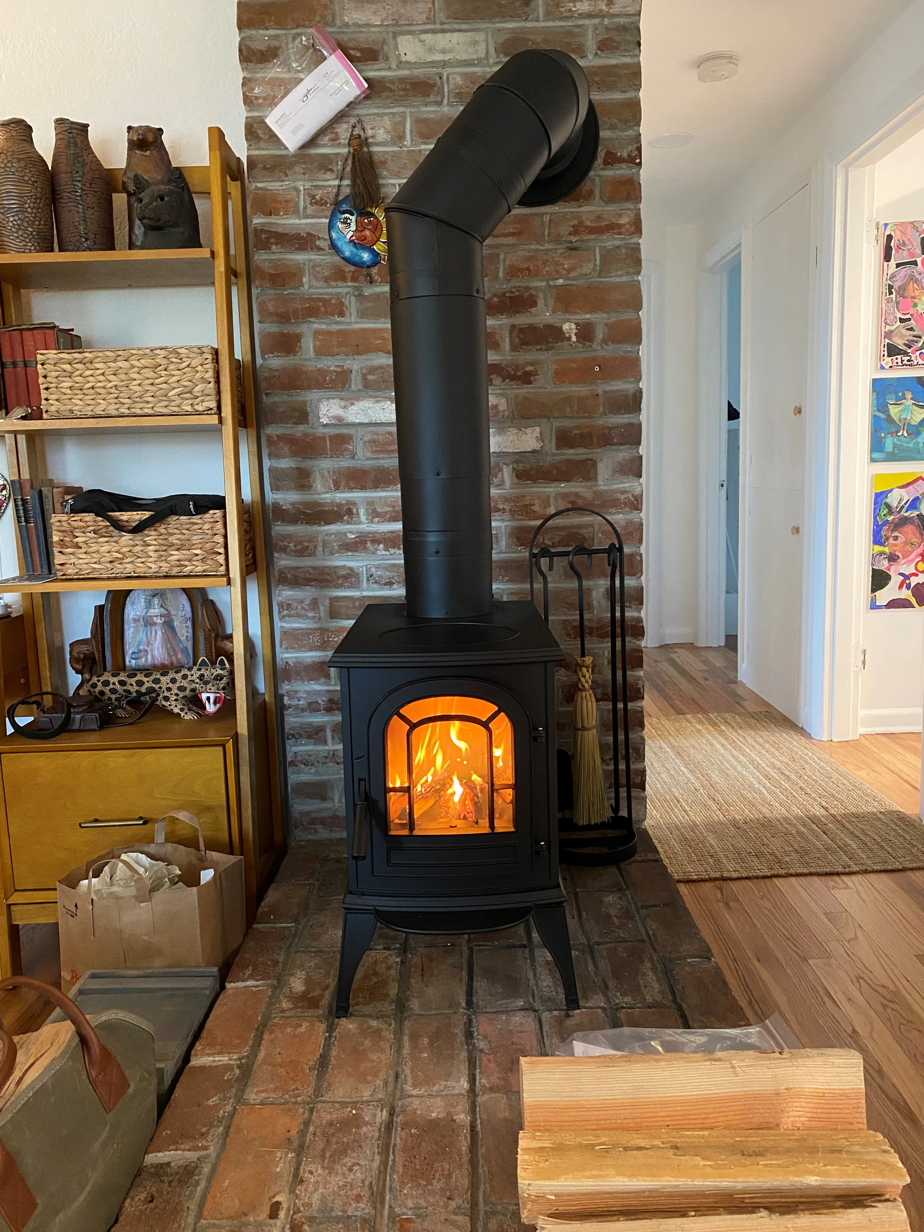 A cozy room with a brick wall and a black wood-burning stove with a fire inside. Firewood and bags are on the floor, and shelves with various items are on the left side.