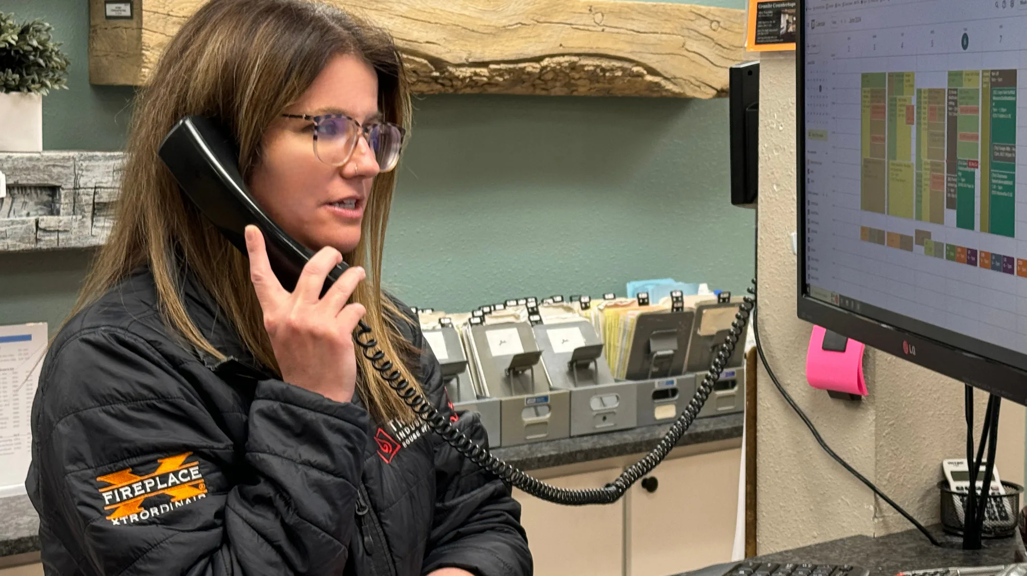 A woman in glasses and a company jacket speaks on a corded phone. She's at an office desk with a wall-mounted calendar and paperwork. A computer screen shows a colorful schedule.