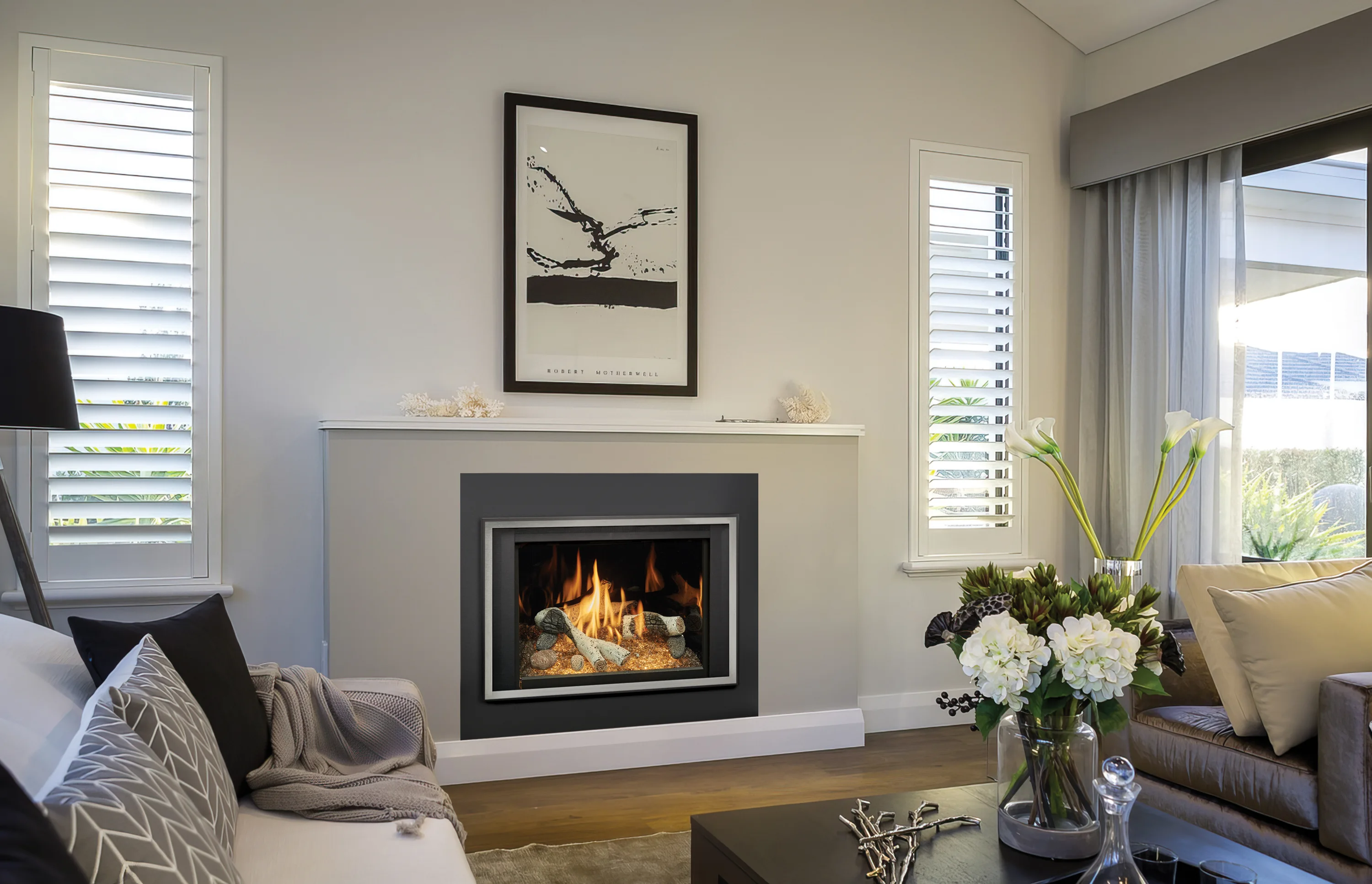 Modern living room with a fireplace, framed art, white couch with gray blanket, and flowers on a coffee table. Shuttered windows and a large glass door light the room.