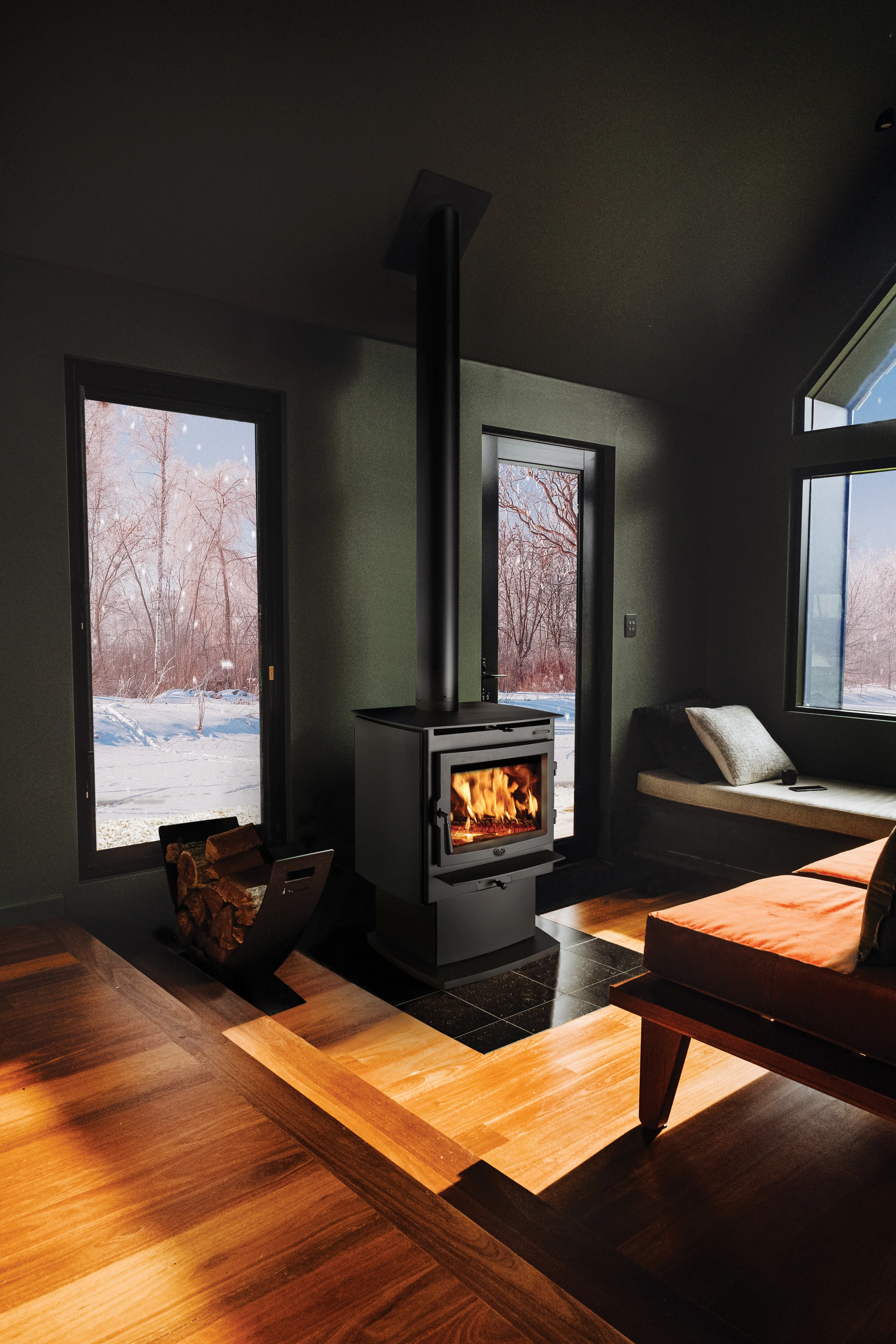 A cozy living room with a wood-burning stove, burning firewood inside. Surrounds include a window, door with outside view of snow-covered trees. Wood basket with logs is on the left.