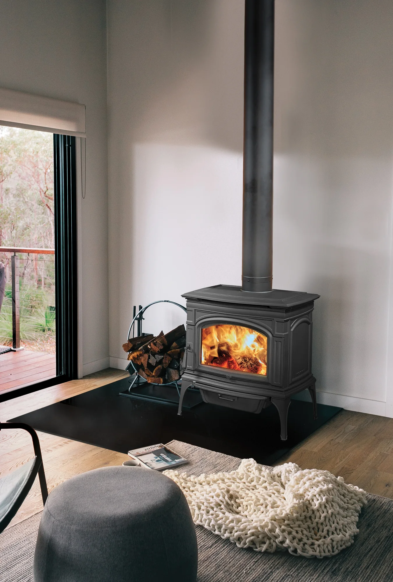 A cozy room with a lit wood-burning stove, firewood in a holder, and a folded cream-colored blanket on a light rug. Sliding glass door opens to an outdoor deck with trees visible.