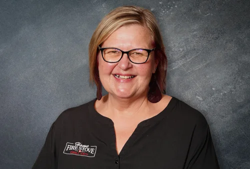 Smiling person with short hair and glasses wearing a black shirt with a logo. The background is a textured gray wall.