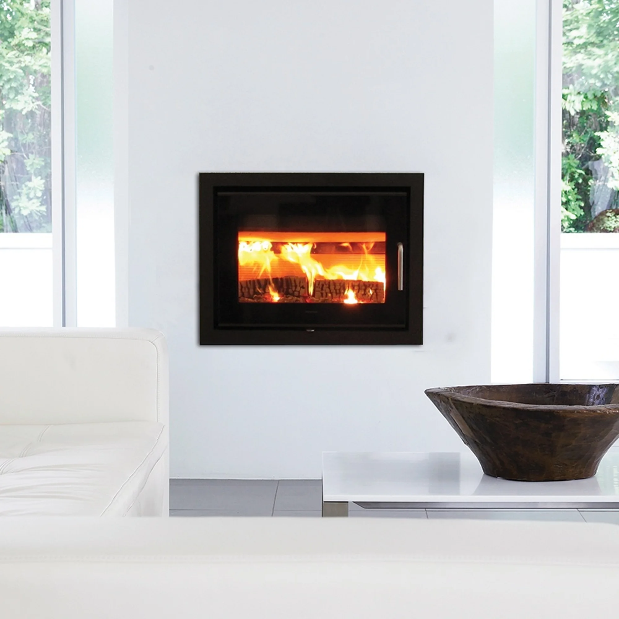 Modern white living room with a black built-in fireplace with visible flames, white leather sofa on left and large wooden decorative bowl on a low white table on right.