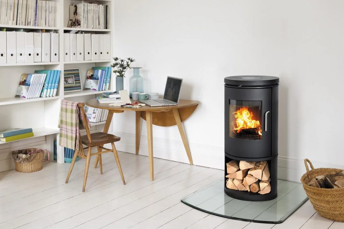 A cozy home office with a wooden chair, a round table, shelves filled with books and binders, a laptop, and other office supplies. A fireplace with burning logs is next to the table.
