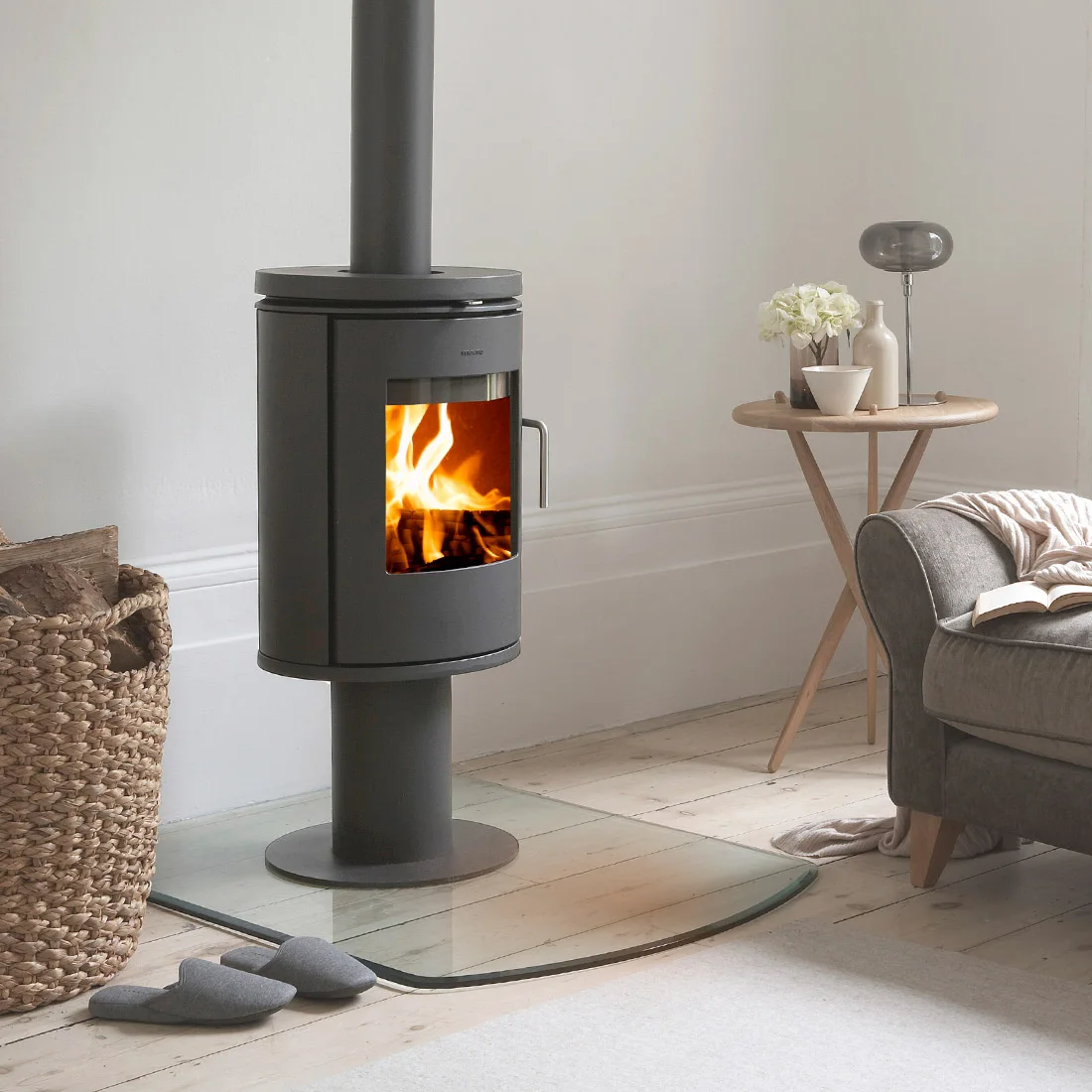 A modern wood-burning stove with a visible fire is set in a cozy living area with a basket of logs, a pair of slippers, a side table with decor, and a gray sofa draped with a blanket.