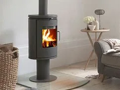 Home office corner with wooden desk and chair, bookshelves, and a modern black wood-burning stove with visible fire and lower log storage on a glass hearth.