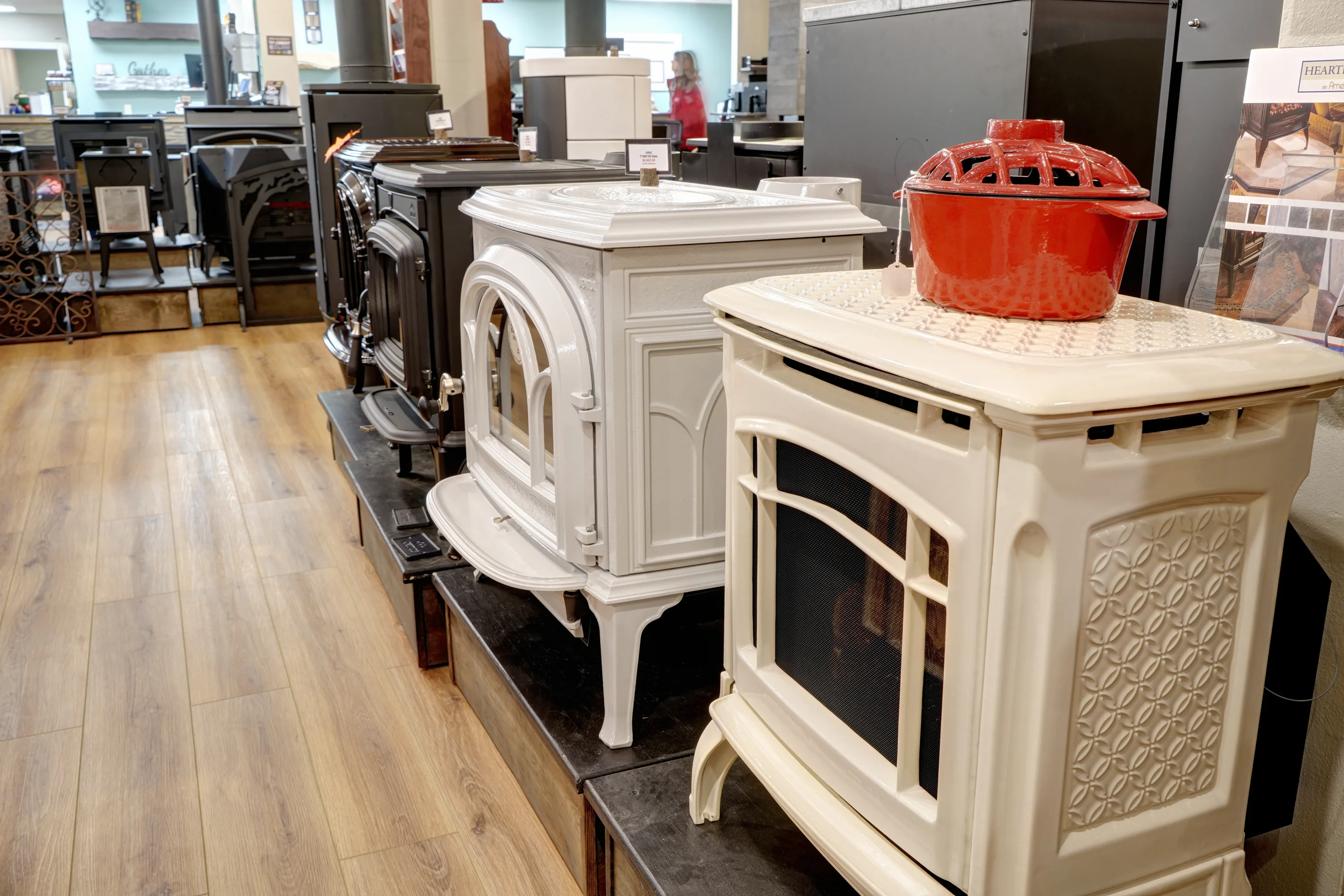 Display of various stove models in a showroom, featuring both traditional and modern designs in black, white, and cream colors, set on wooden and platform floors.