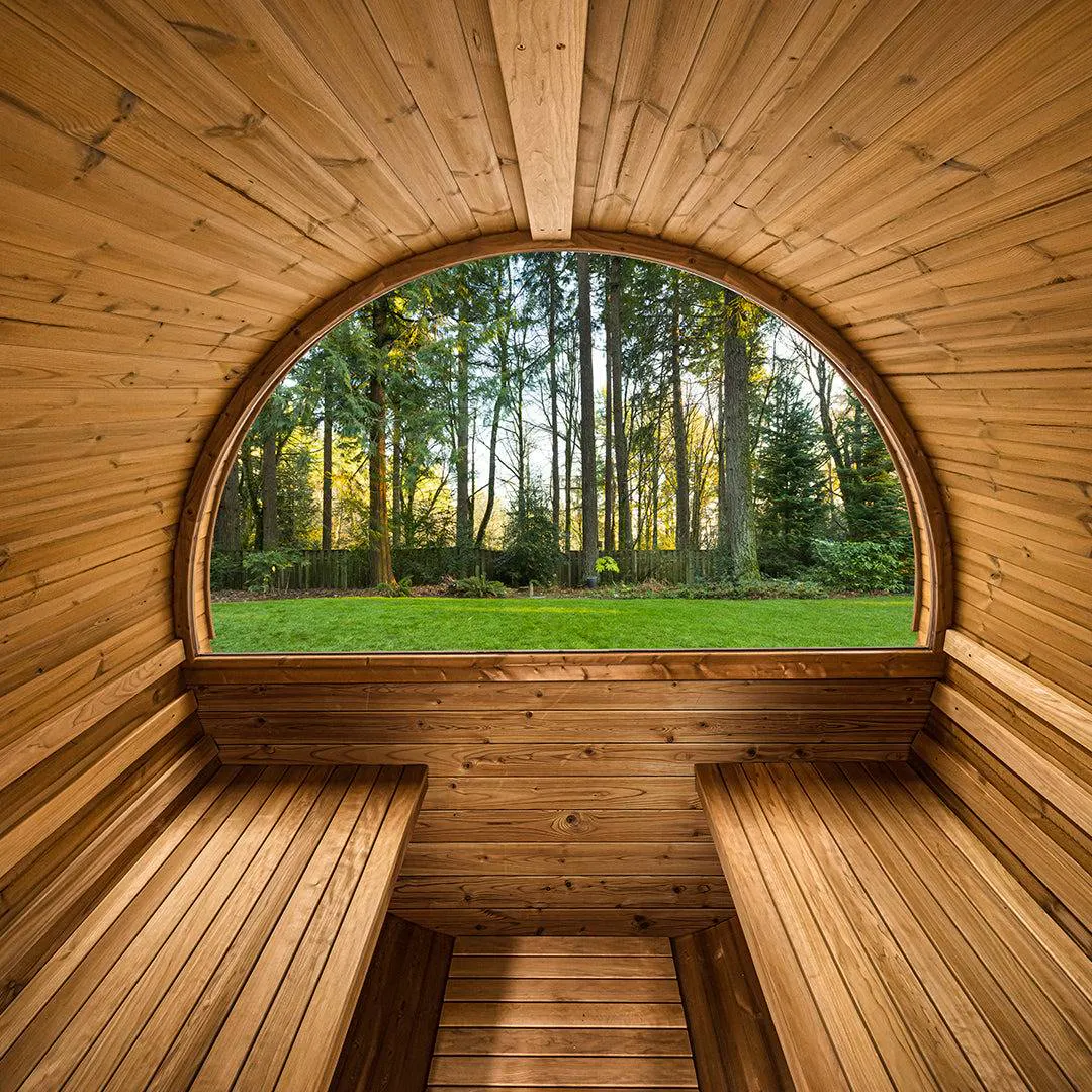 Interior of a wooden barrel sauna with bench seating, looking out a rounded window at a green lawn and trees in the background. Sunlight illuminates the peaceful outdoor scenery.