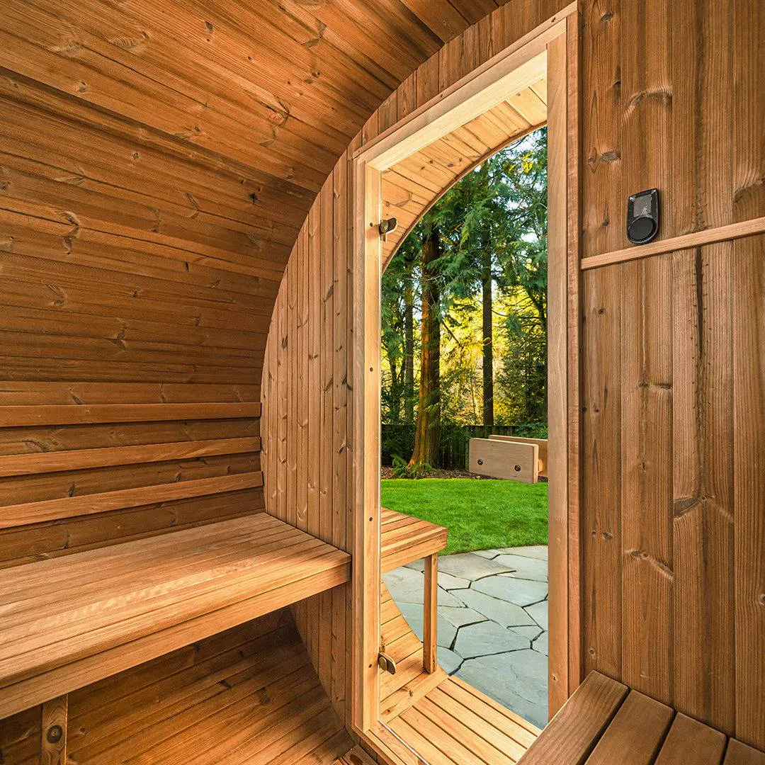 Inside view of a wooden sauna with benches, looking out through an open door to a sunny backyard with green grass, stone pathway, and trees.