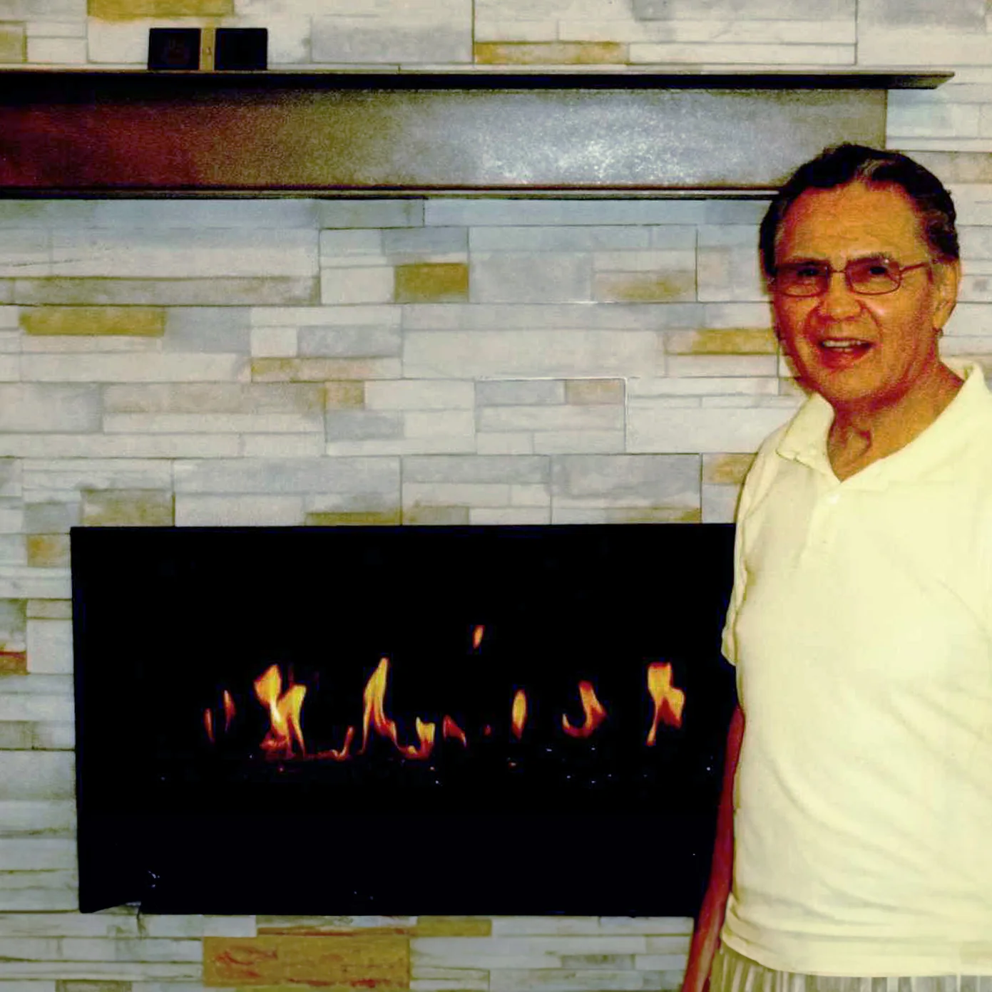 Man in a light shirt standing beside a modern stone fireplace with visible flames. A dark mantel holds small decorative items above the fireplace.
