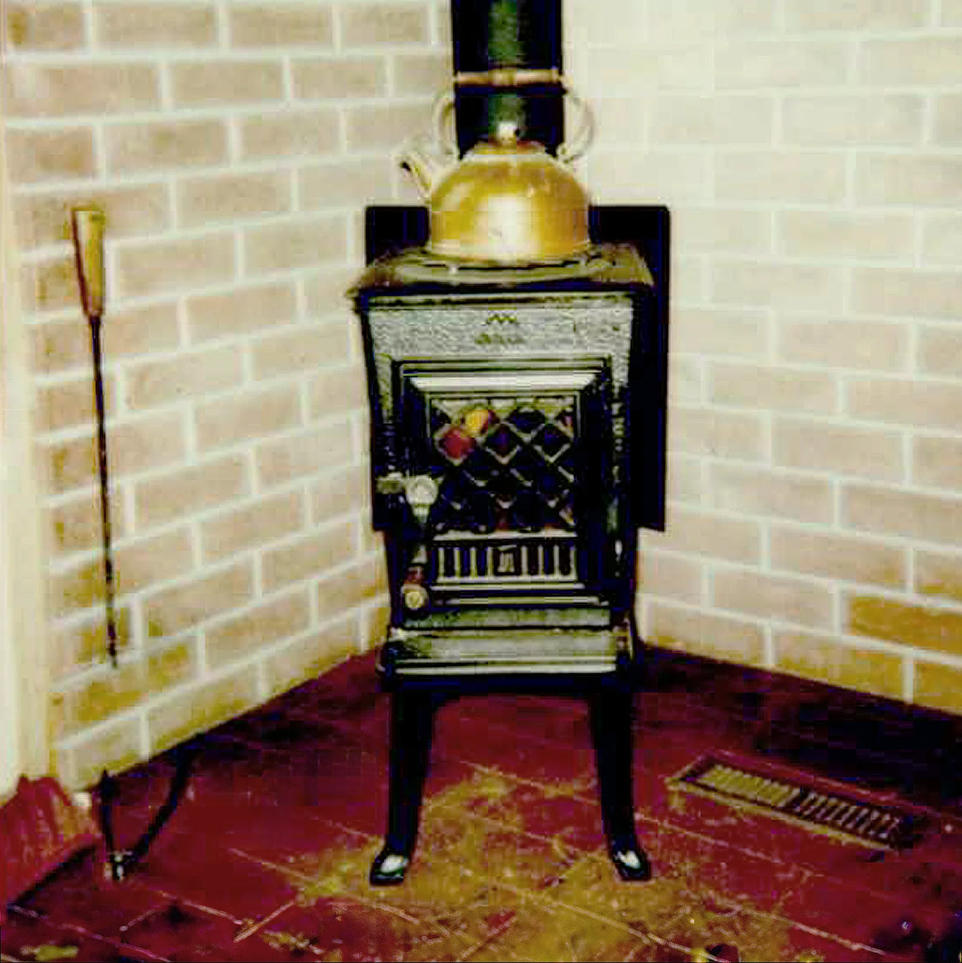 A black cast iron stove with intricate detailing sits in a brick corner. A brass kettle rests on top. A fireplace tool is on the left, and a floor vent is on the right.