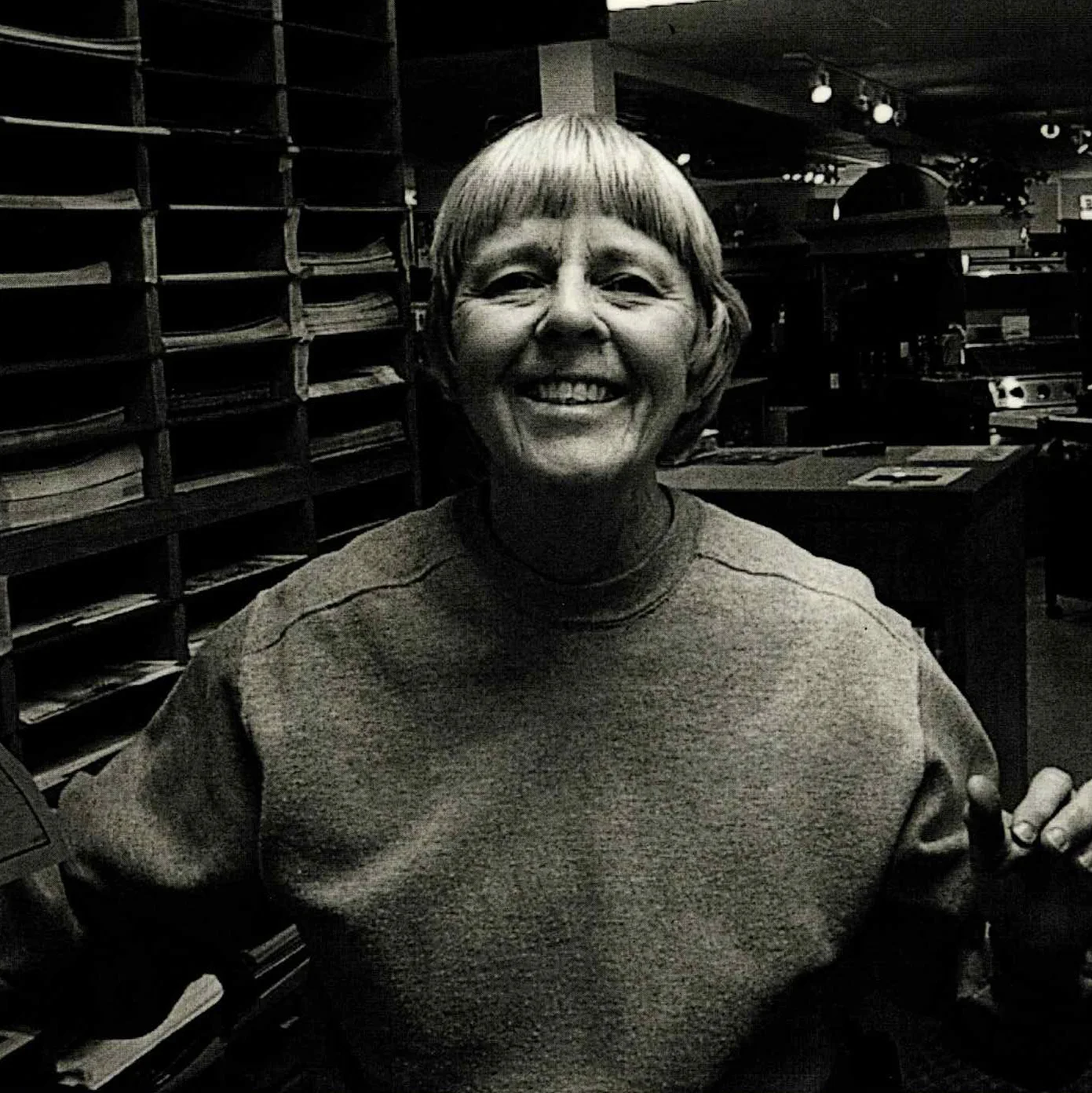 A person with short hair smiling broadly, wearing a sweatshirt in a room with shelves filled with papers and a dimly lit background.