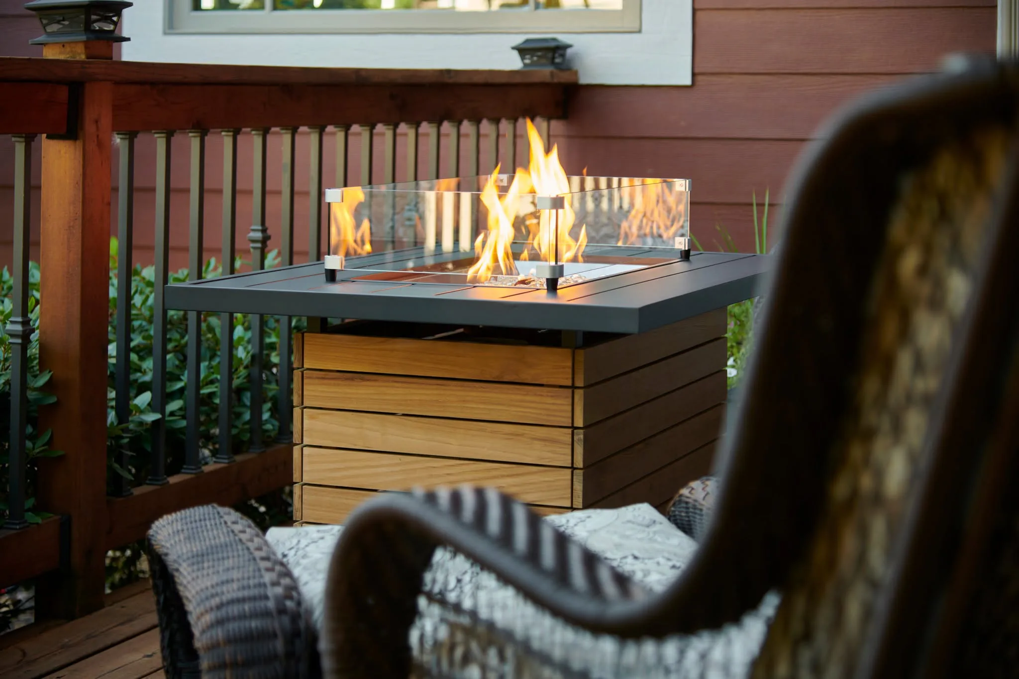 Rectangular outdoor fire pit table with glass wind guard and flames on a wooden deck, wicker chairs and railing nearby