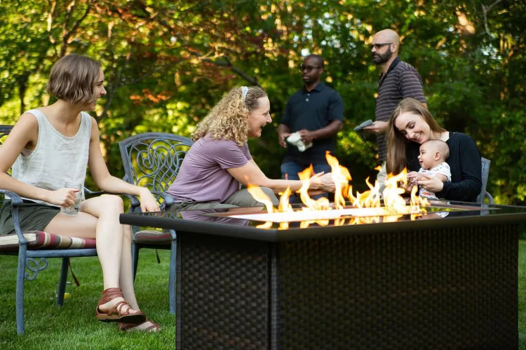 Adults and a mother holding a baby seated around a lit outdoor fire pit in a backyard