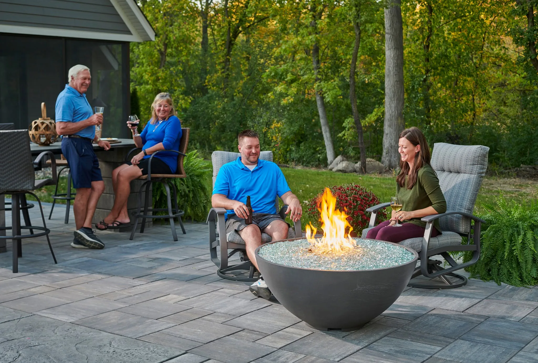 Four adults socializing around a backyard fire pit on a paved patio, holding drinks with trees in the background