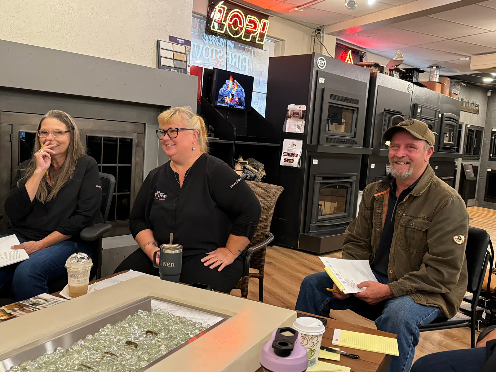 Three people sit around a table in a store with fireplaces and stoves. They are smiling, holding papers, and have drinks on the table. Neon signs are visible in the background.