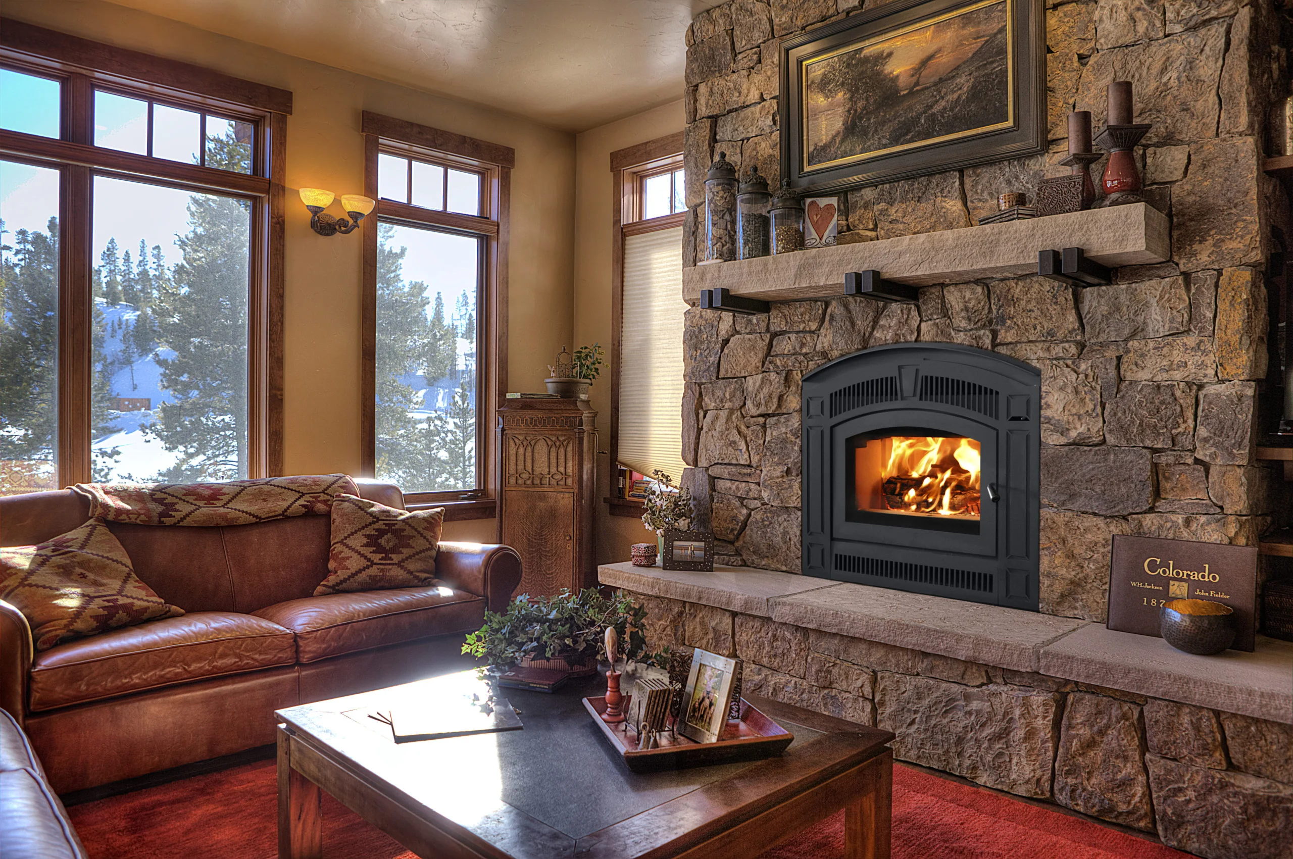Cozy living room with large windows, a leather sofa with patterned cushions, wooden furniture, a stone fireplace with a burning fire, framed art, and a coffee table with decor items.