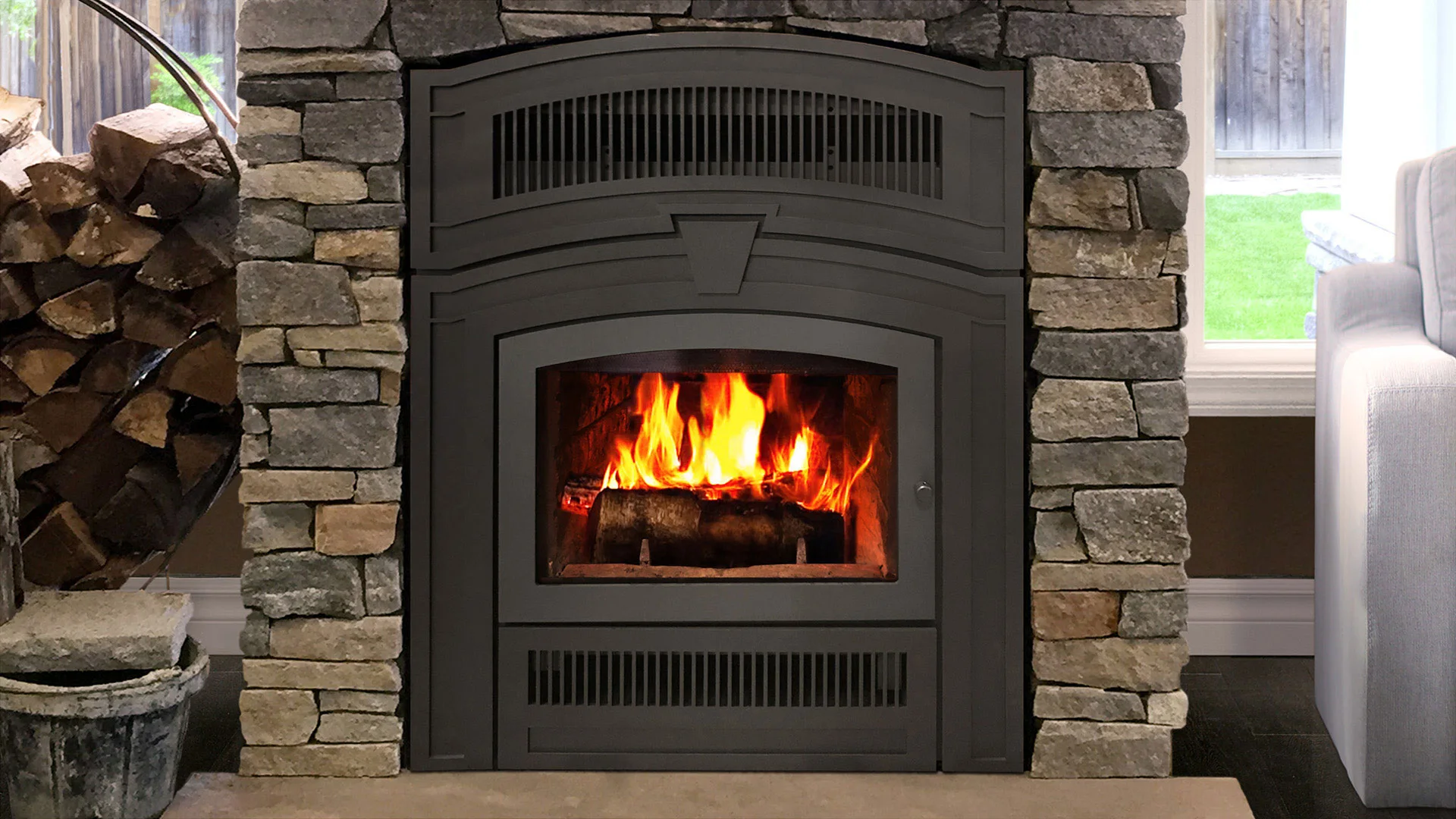 A stone fireplace with a metal insert and a blazing fire inside. To the left, a stack of firewood is visible. A window and part of a couch are seen on the right side.