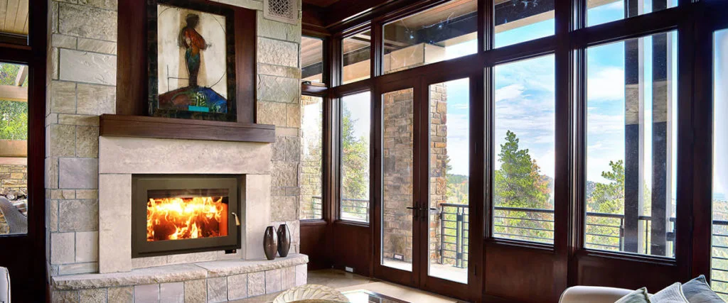 Modern living room with large windows and a stone fireplace. A warm fire is burning, and a painting hangs above the mantle. Outside, trees and a balcony are visible through the windows.