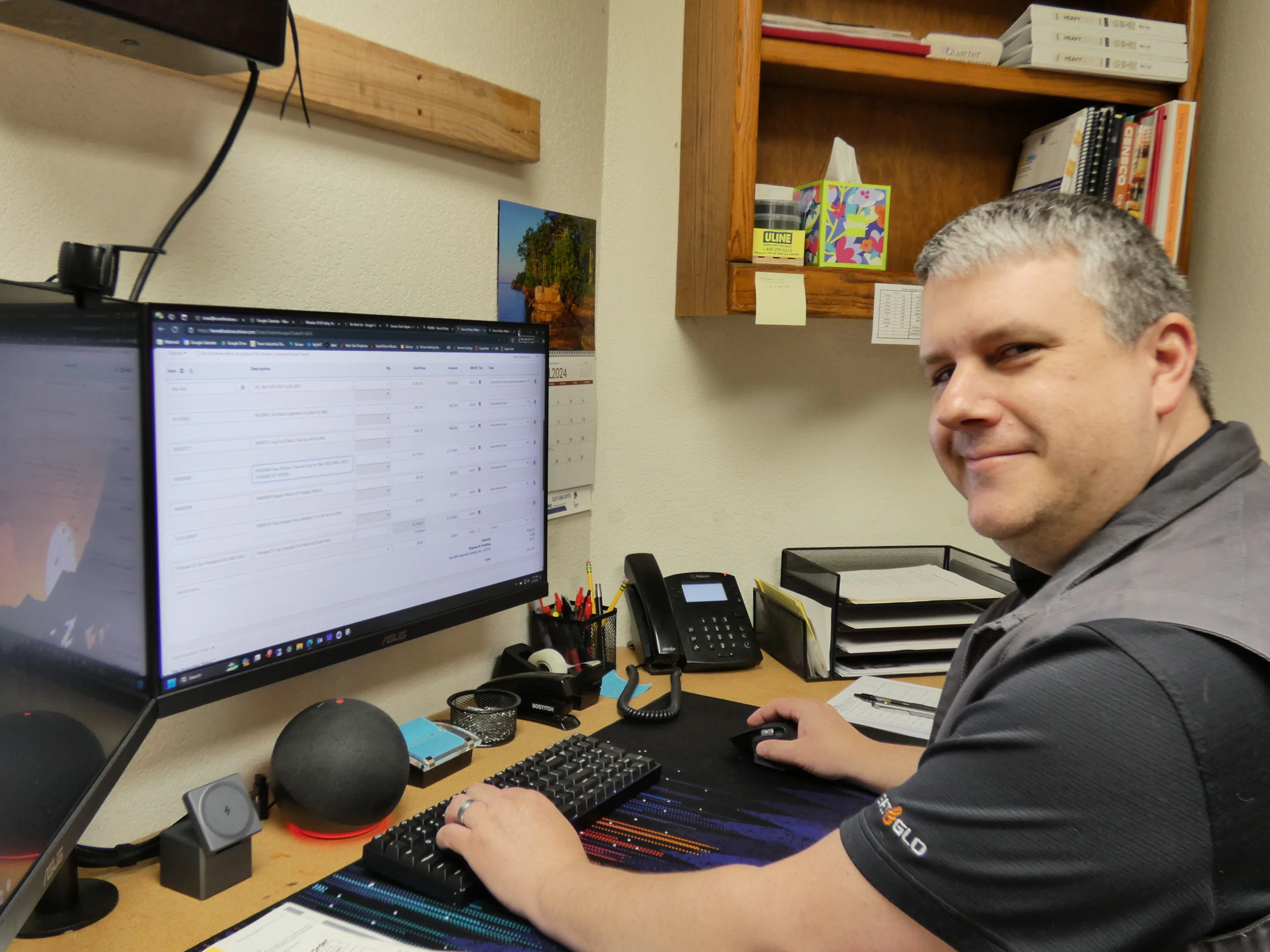 Smiling man at desk using mouse and keyboard beside dual monitors, desk phone, pens and shelves with tissue box and books.