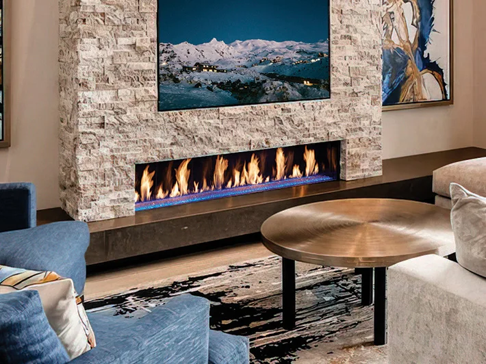 Contemporary living room with long built-in linear fireplace in a stone wall beneath a mounted TV, round brass coffee table, blue armchair, beige sofa and abstract rug.