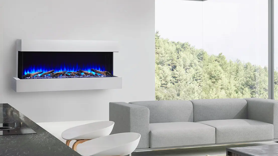 Modern living room with a gray sectional sofa, a wall-mounted electric fireplace emitting blue flames, two white chairs, and large windows with a view of a forest outside.