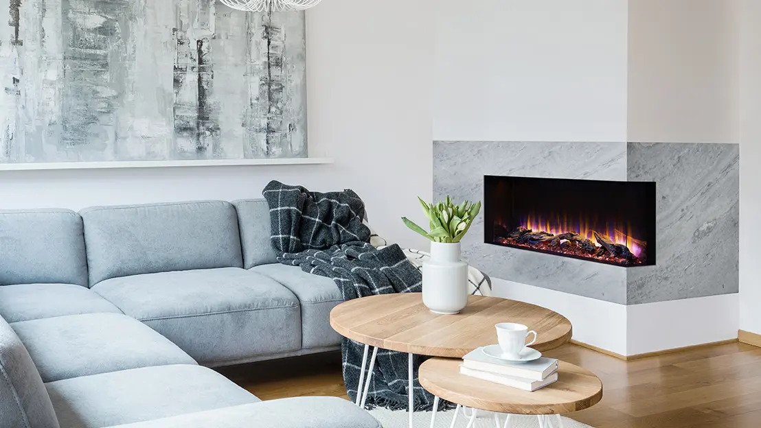 Living room with a light-gray sectional sofa beside a modern electric fireplace, gray and black blanket, abstract painting, wooden coffee table with a plant, and a cup on a saucer.