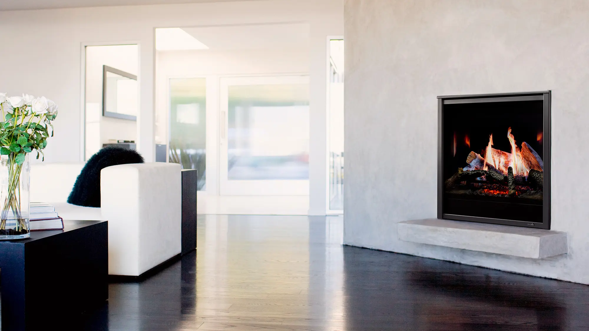 Modern living room with a white couch, black coffee table with a flower vase, dark wooden floor, and a wall-mounted fireplace with burning logs, creating a cozy ambiance.