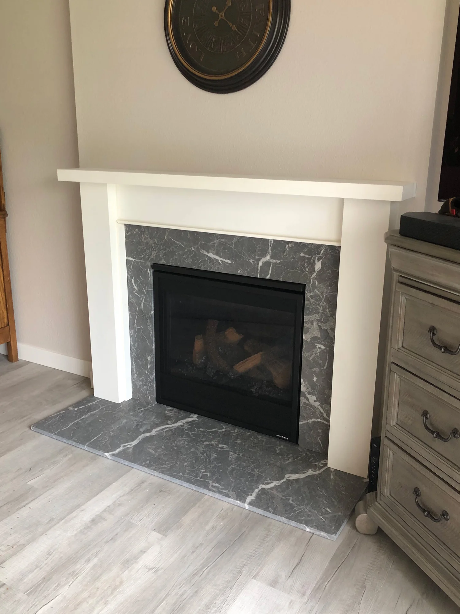 A marble fireplace with a white mantel, situated in a living room with light-colored wood flooring. A round, dark-framed clock hangs above the fireplace.
