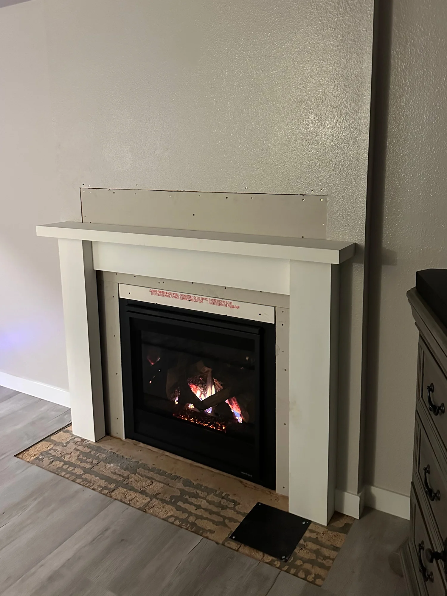 A lit gas fireplace with a white mantle under construction is attached to a beige wall. The fireplace sits on a partially finished floor.